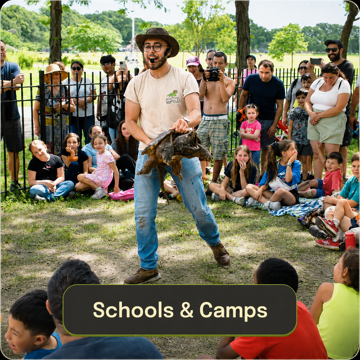 A man performing a reptile demonstration in long island holding a large turtle in front of a crowd of children and adults at an outdoor park, with trees and grassy area in the background and a sign that reads 'Schools & Camps'.