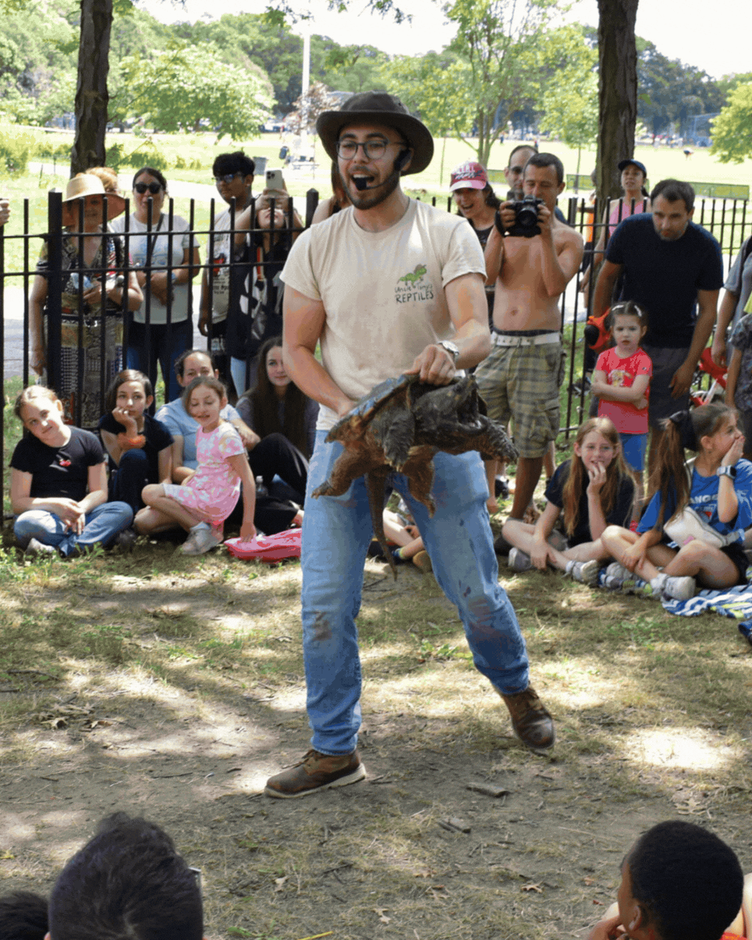 A man wearing a safari hat and glasses is holding a small crocodile during a wildlife demonstration, with children and adults sitting and standing around him, watching attentively in an outdoor park setting.