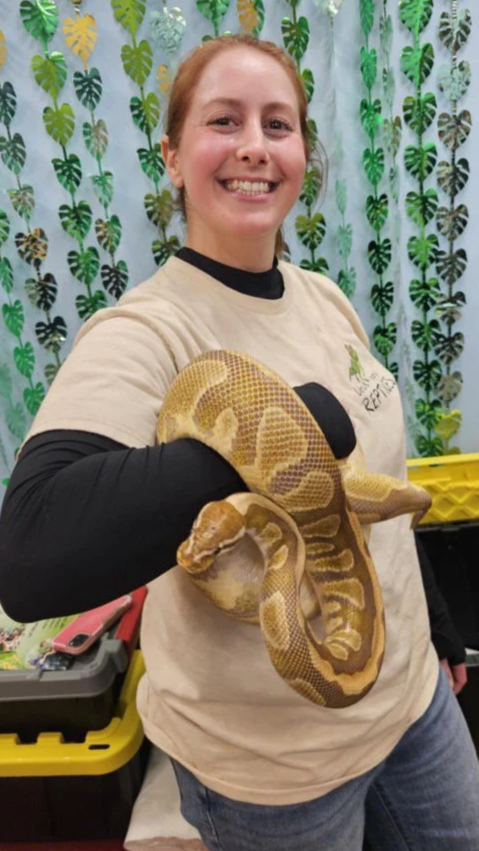 A woman smiling and holding a large yellow and brown ball python snake in her arms, standing in front of a green leaf-patterned backdrop at a birthday party