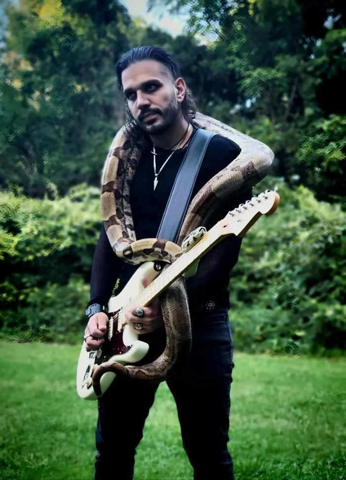 A man with long dark hair, wearing a black shirt and dark jeans, stands outdoors holding an electric guitar. A large snake is draped around his shoulders and neck, with its body wrapped around his arm and guitar. The background includes trees and green foliage.