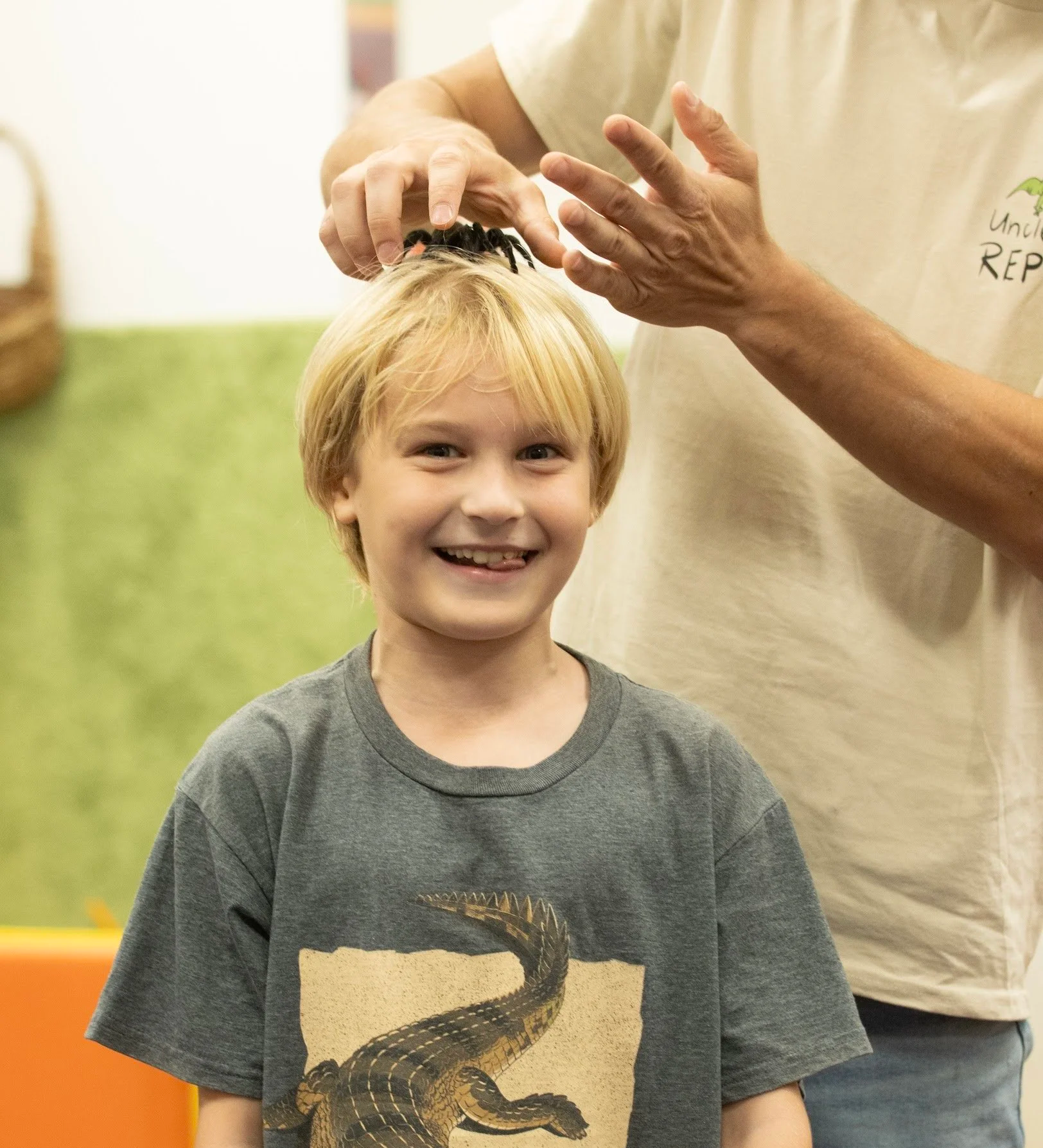 A young blonde-haired boy smiling, wearing a dark gray T-shirt with a crocodile design, standing indoors at an event with a person in a beige shirt nearby. it is a reptile show at a school