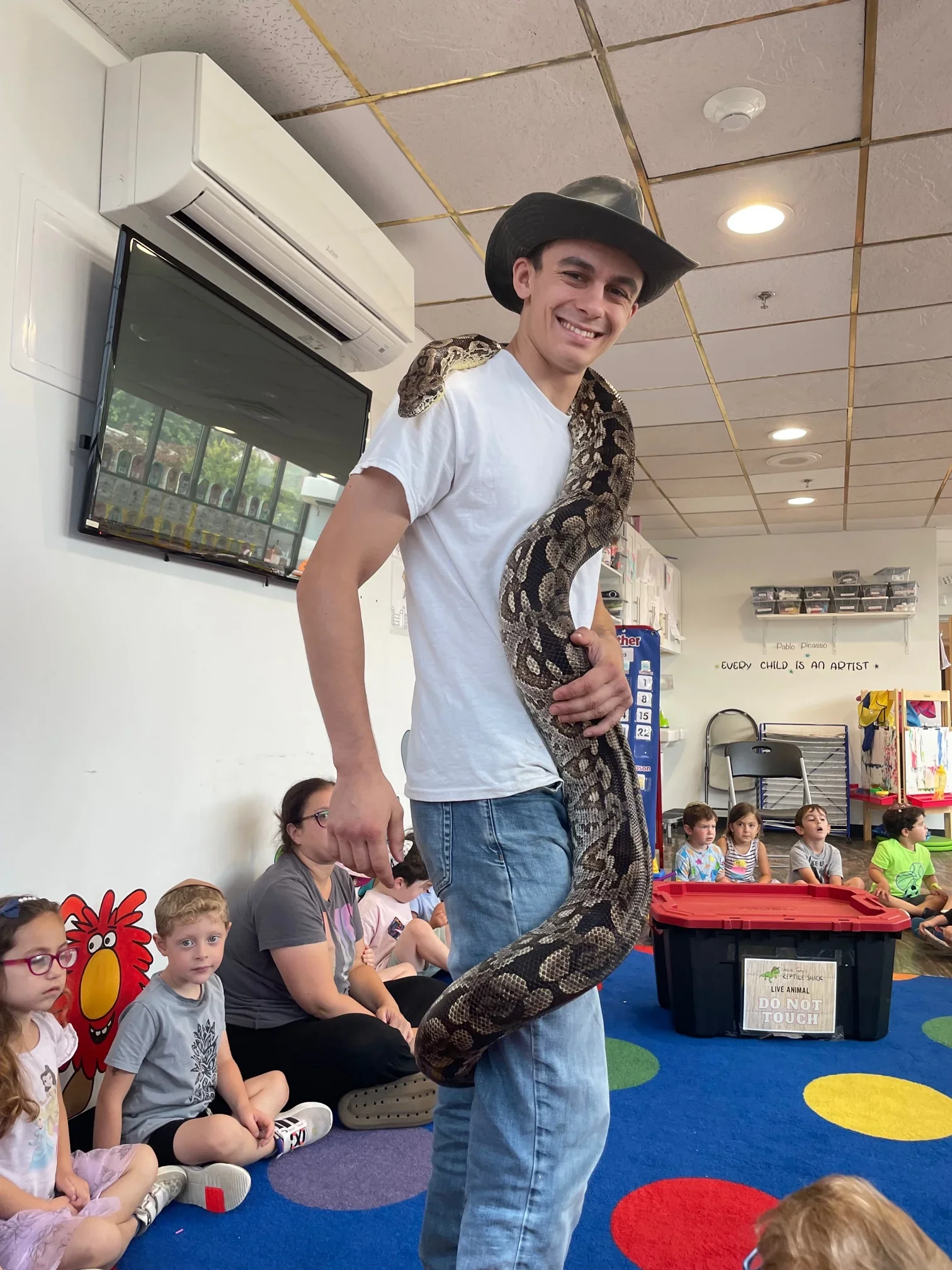 A young man wearing a cowboy hat and white t-shirt, smiling, holding a large snake around his neck, in a classroom with children sitting on the floor.