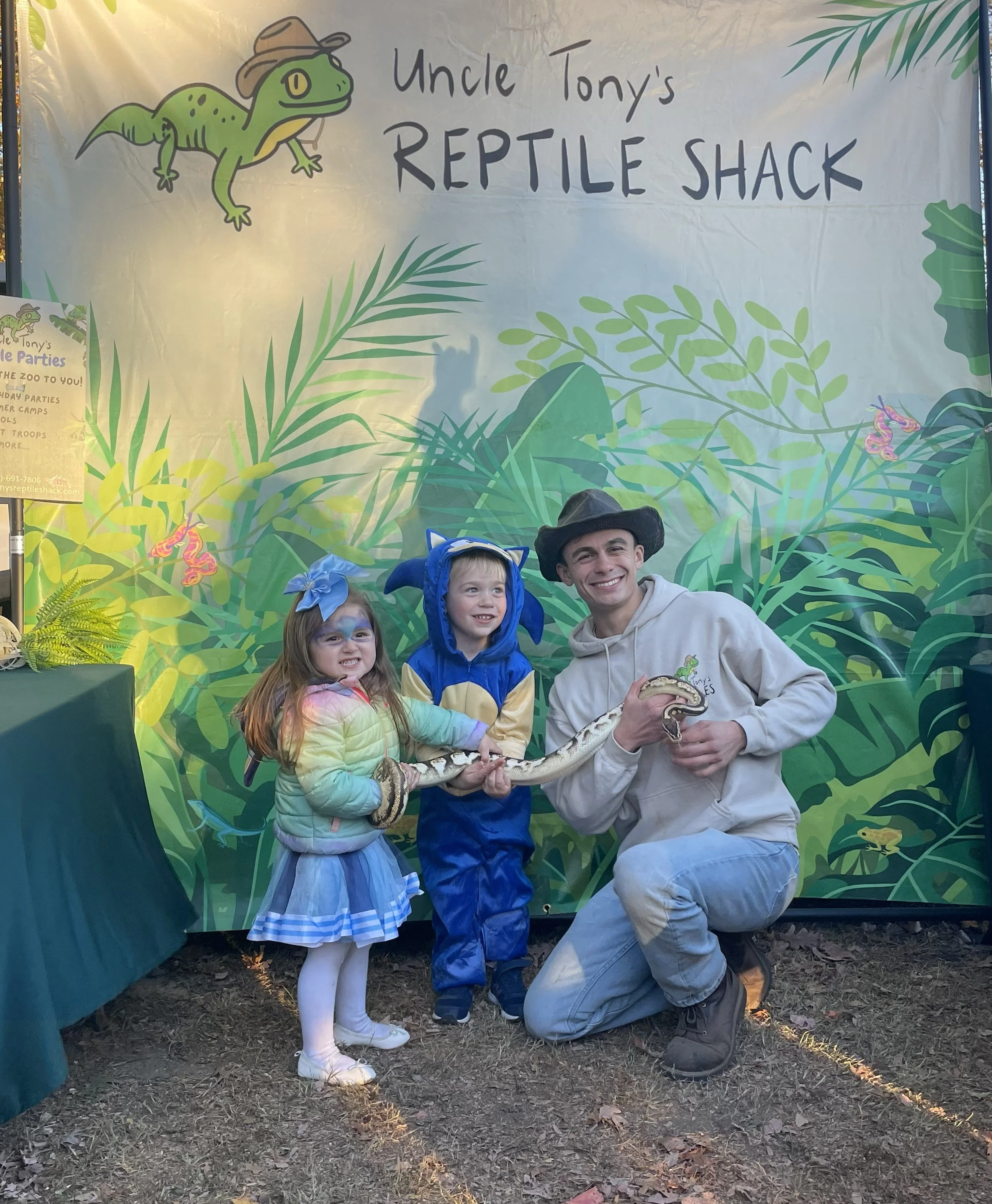 A group of four people, including two children and two adults, is posing for a photo at Uncle Tony's Reptile Shack. The children are dressed in colorful outfits, one with face paint and the other in a blue and yellow costume. One adult is kneeling, holding a snake, and the other is standing behind them. The background features a green leafy jungle scene and a sign with the name of the reptile shack and a cartoon iguana wearing a hat.