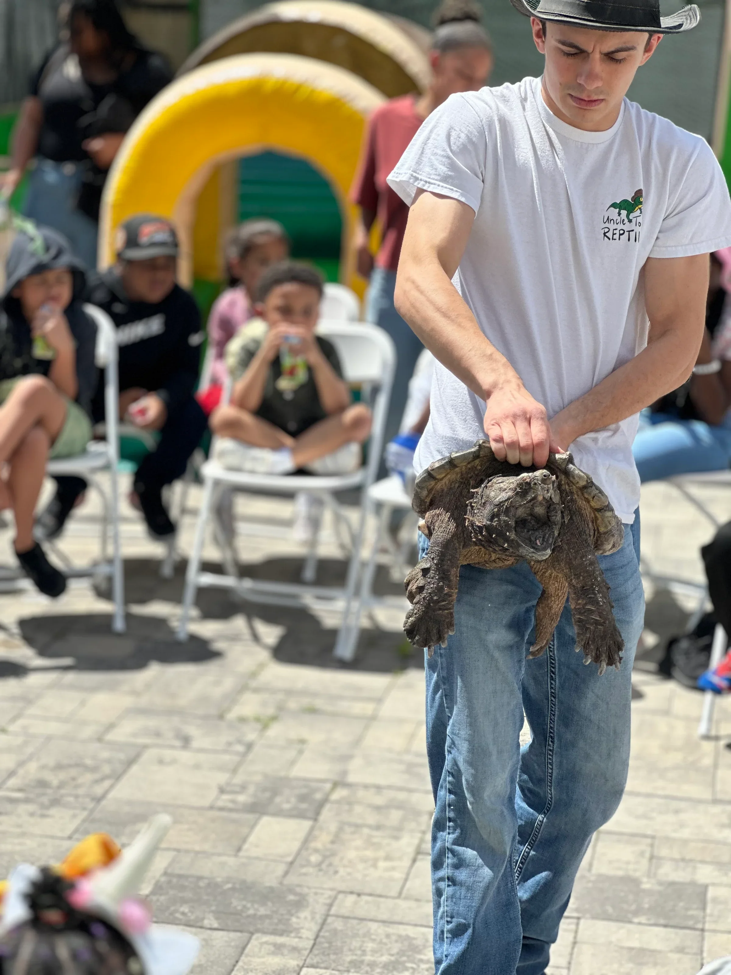 A young man wearing a white t-shirt with a dinosaur logo and the text 'Uncle T to Rept', blue jeans, and a western-style hat is holding a large turtle. In the background, there are several seated children and adults watching, and a colorful yellow and green arch structure.