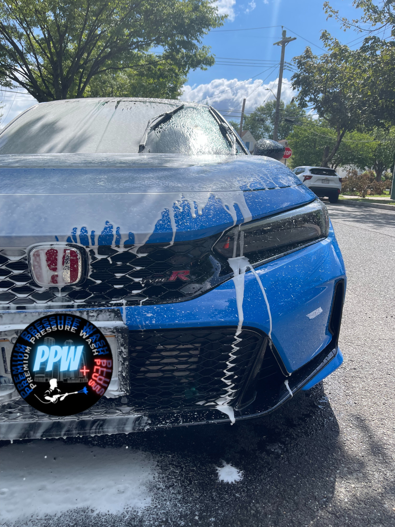 Blue Honda Civic Type R being washed with soap and water, foam dripping down the front of the vehicle, parked outdoors on a sunny day with trees, houses, and other cars in the background.