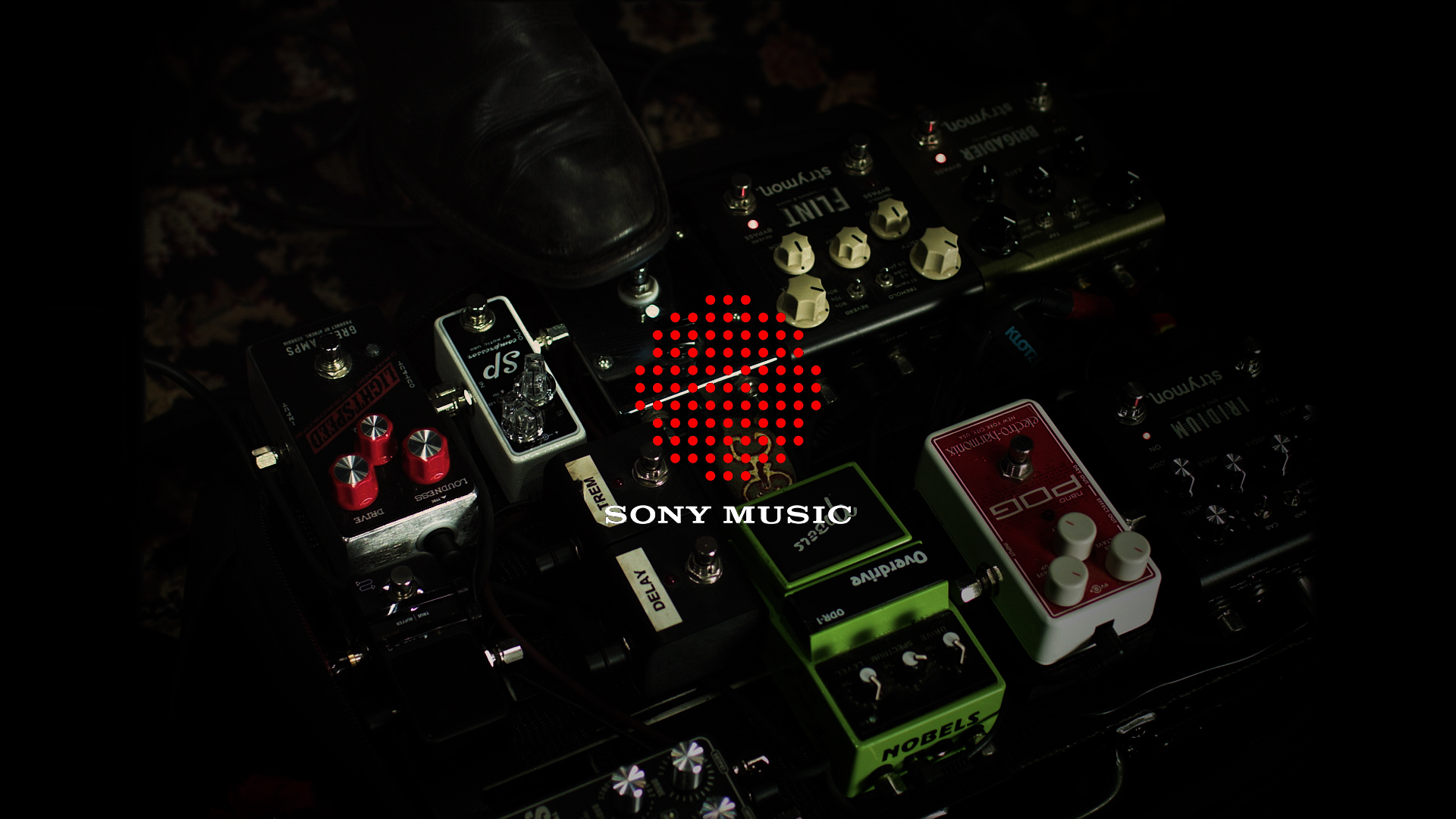 Close-up of a guitar pedalboard with various effects pedals, a shoe resting on top, and a Sony Music logo overlay.
