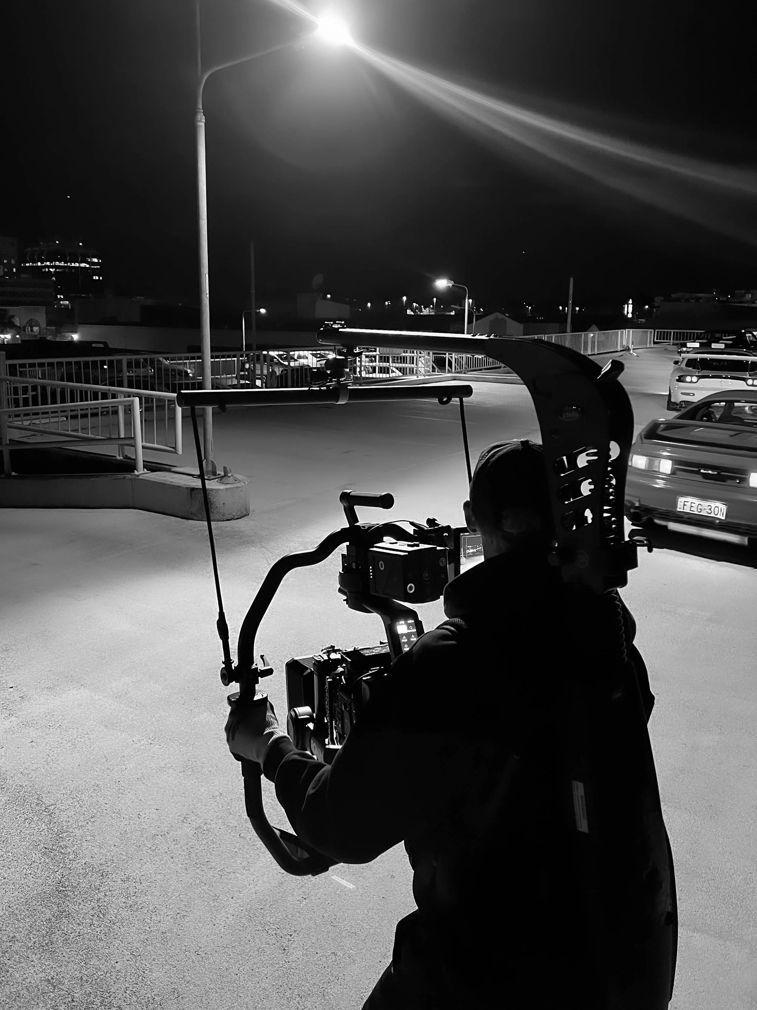 A person operating a camera stabilizer on a rooftop parking lot at night, with a streetlight illuminating the scene.