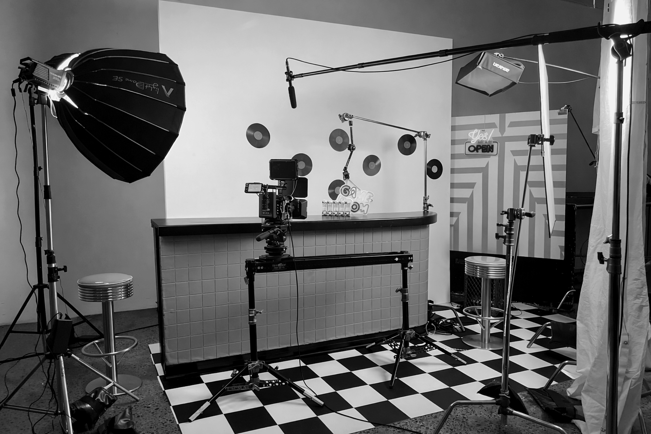 Photography studio set with lighting equipment, camera, and backdrops. Black and white interior with checkered floor and various studio tools arranged around a counter.