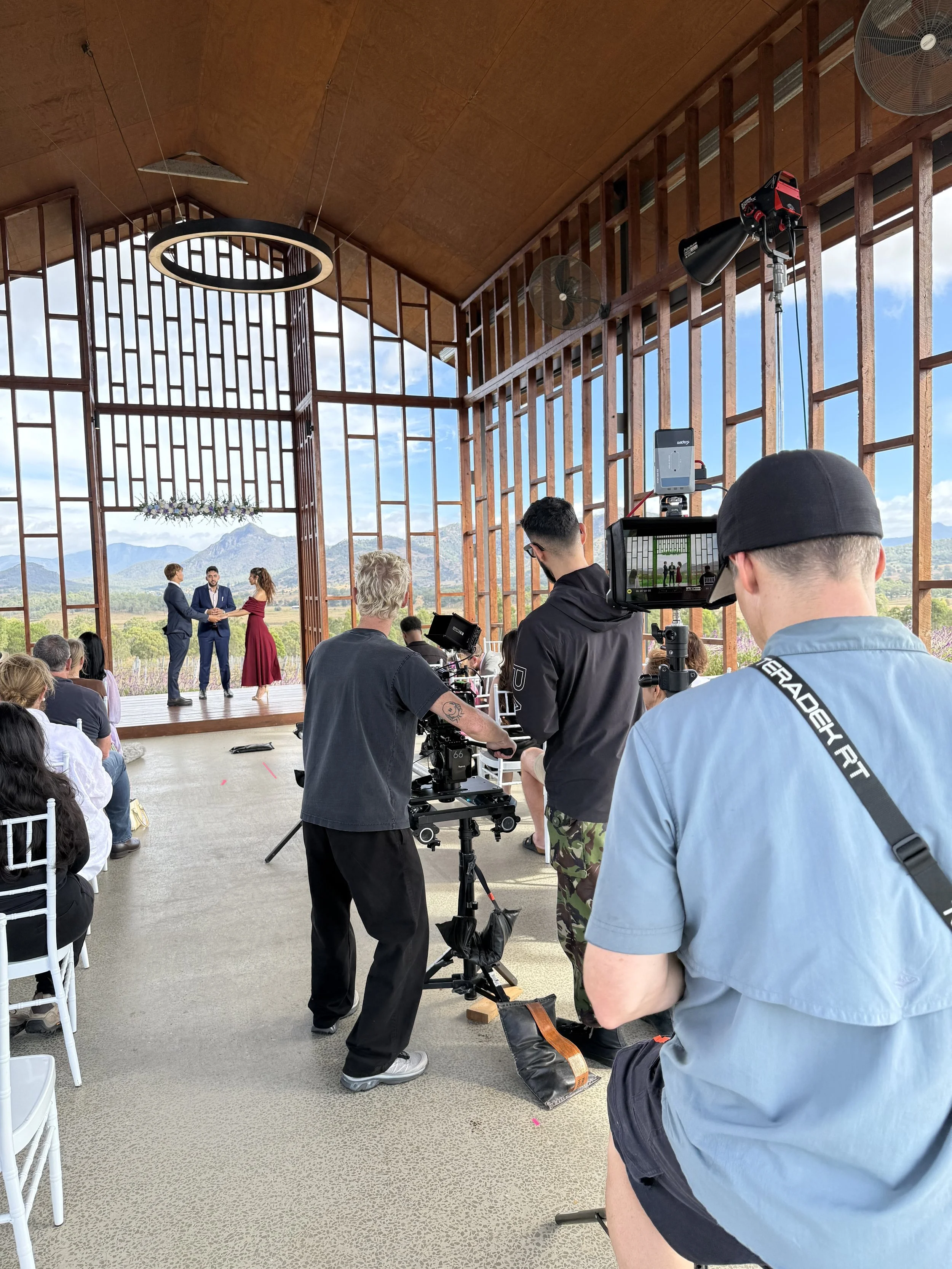 A wedding ceremony taking place in a venue with open sides, mountains in the background, and a group of people filming the event with professional equipment. The couple is standing with an officiant in between them, holding hands.