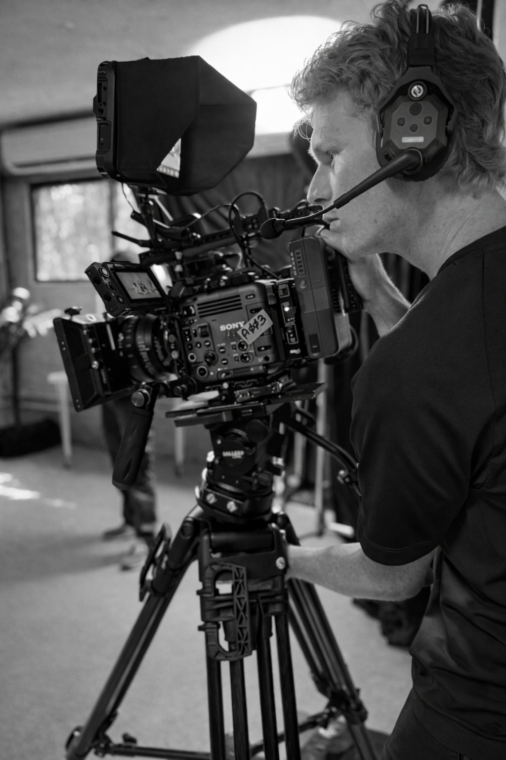 A man with curly hair wearing headphones operating a professional Sony video camera on a tripod in a room with a window.