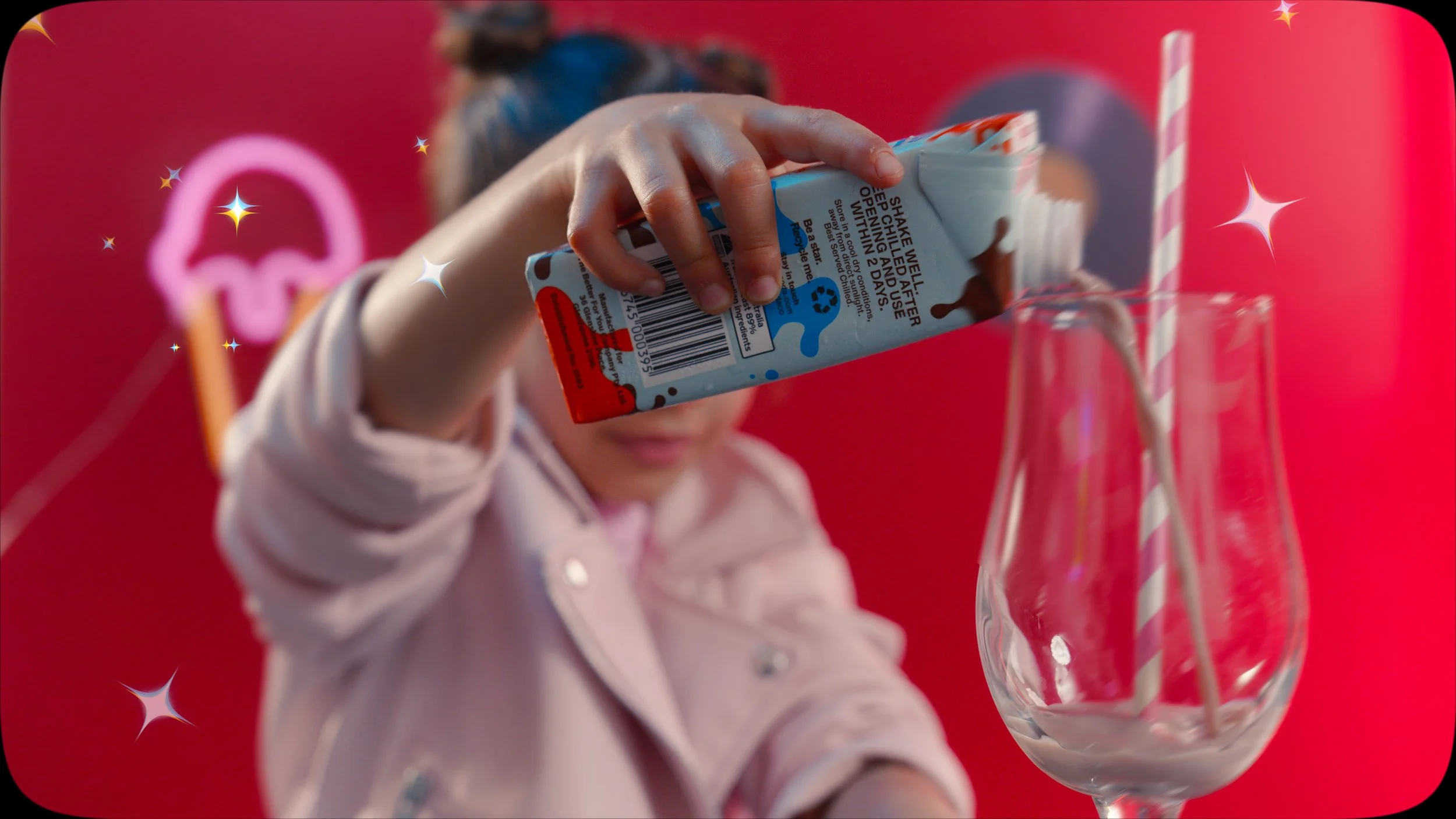 A young girl in a pink jacket pouring milk from a carton into a glass with a striped straw, against a red background with a neon pink umbrella decoration.