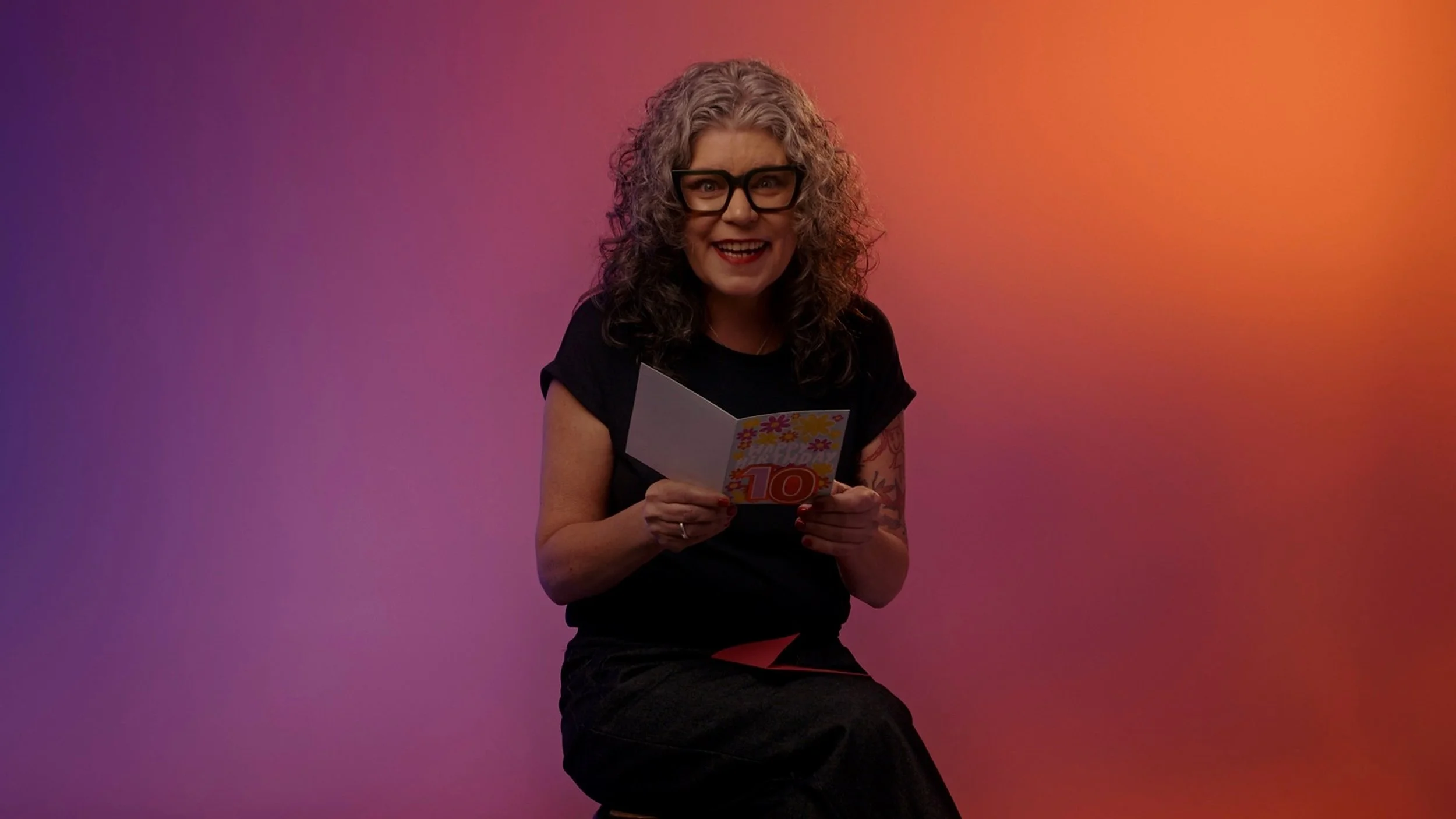 A woman with curly hair and glasses smiling while holding a colorful card, sitting against a vibrant gradient background.