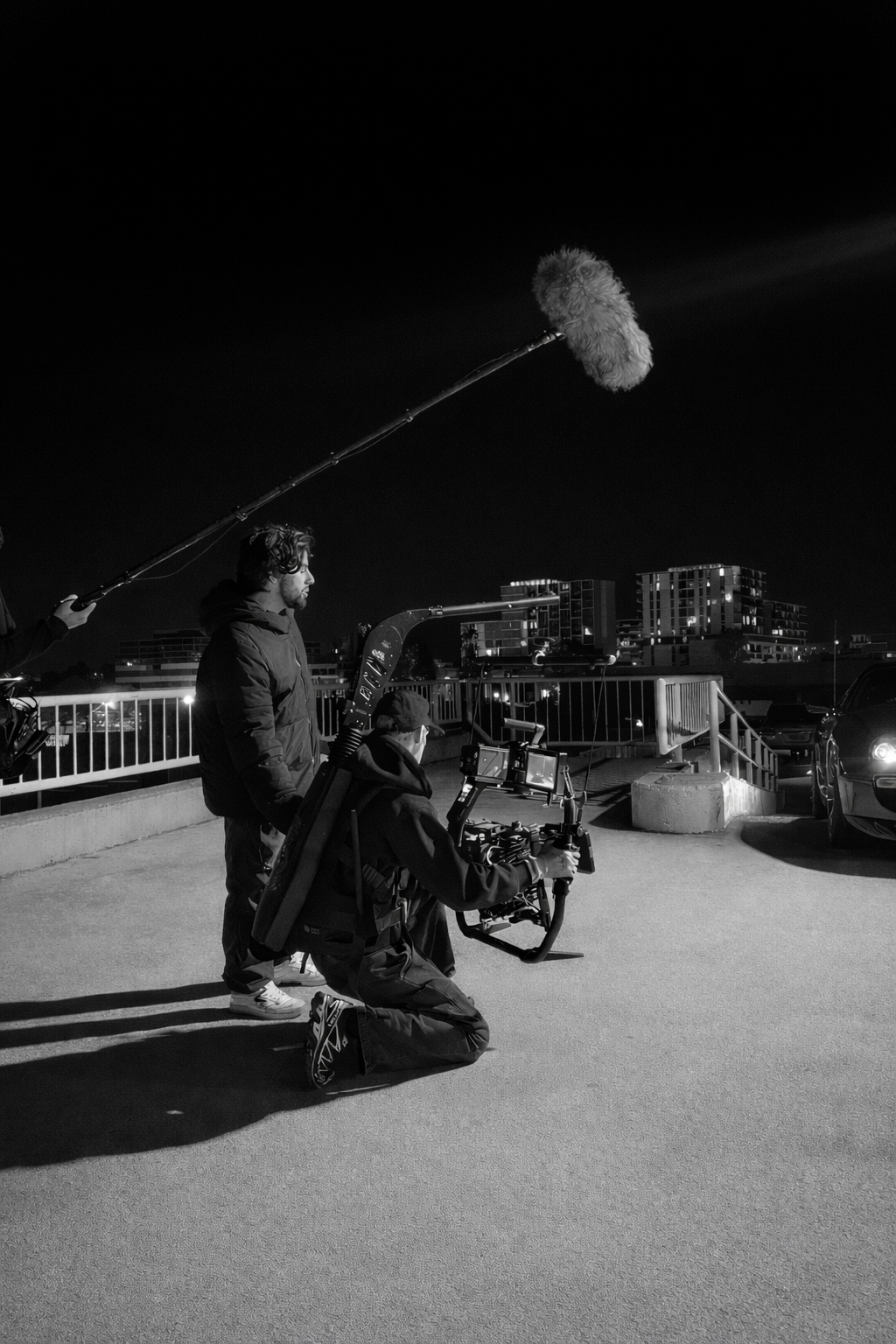 Black and white photo of a film crew filming outdoors at night, with a camera operator kneeling and a man standing beside him, holding a boom microphone with a furry windscreen, with buildings in the background and parked cars nearby.