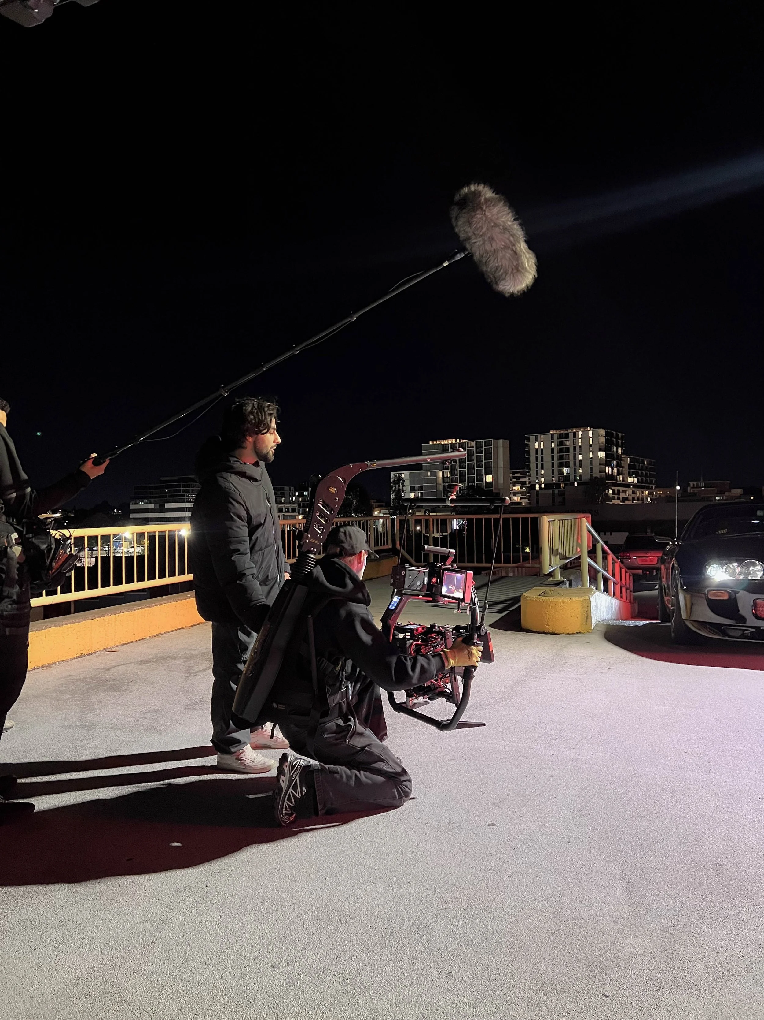 Filming crew capturing a scene on a rooftop parking lot at night, with city buildings in the background.