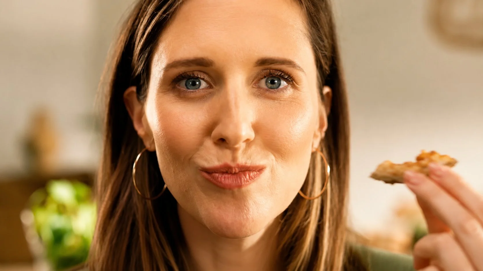 A woman with brown hair and hoop earrings holding a slice of pie close to her mouth, smiling softly at the camera.