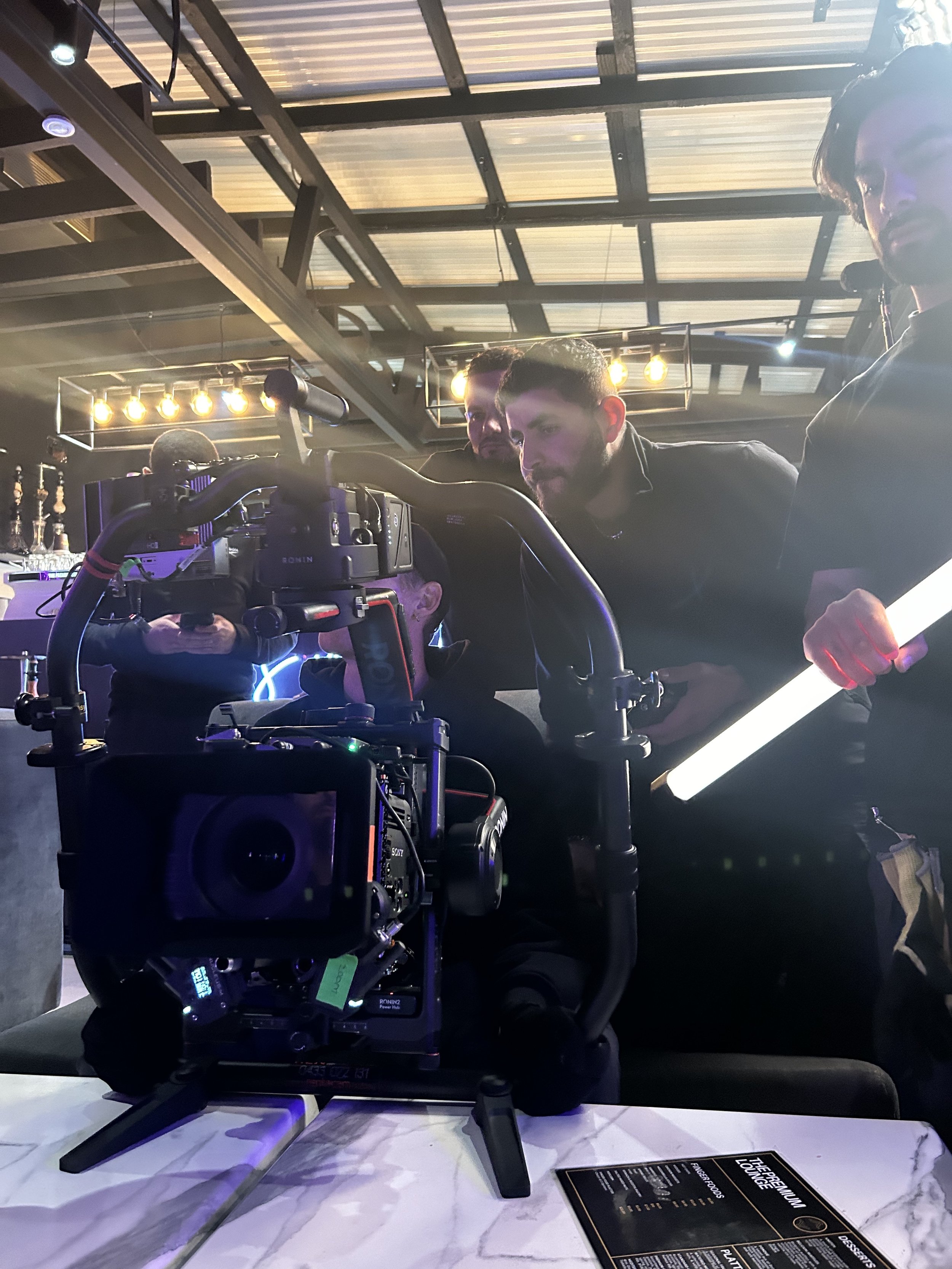 Close-up of a camera on a tripod capturing a scene, with two men observing the shot in an indoor setting with industrial ceiling lighting.