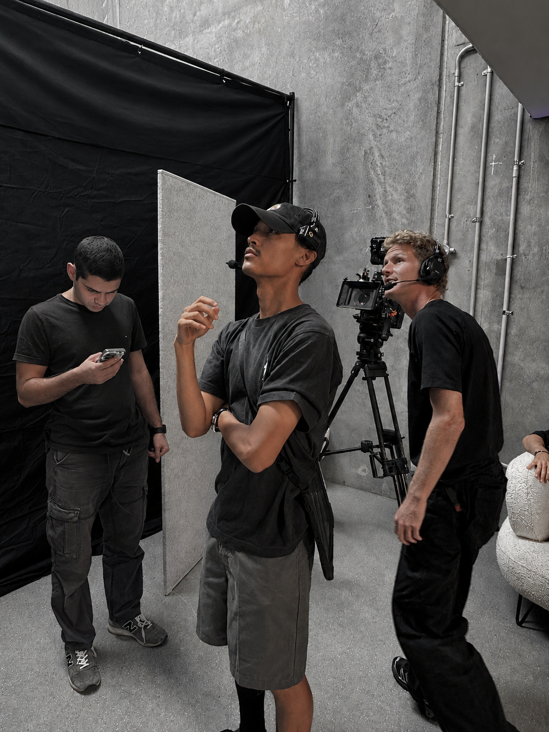 Three men in a production studio; one is looking at his phone, another is standing with a headset and pointing upward, and the third is filming with a camera while wearing a headset.