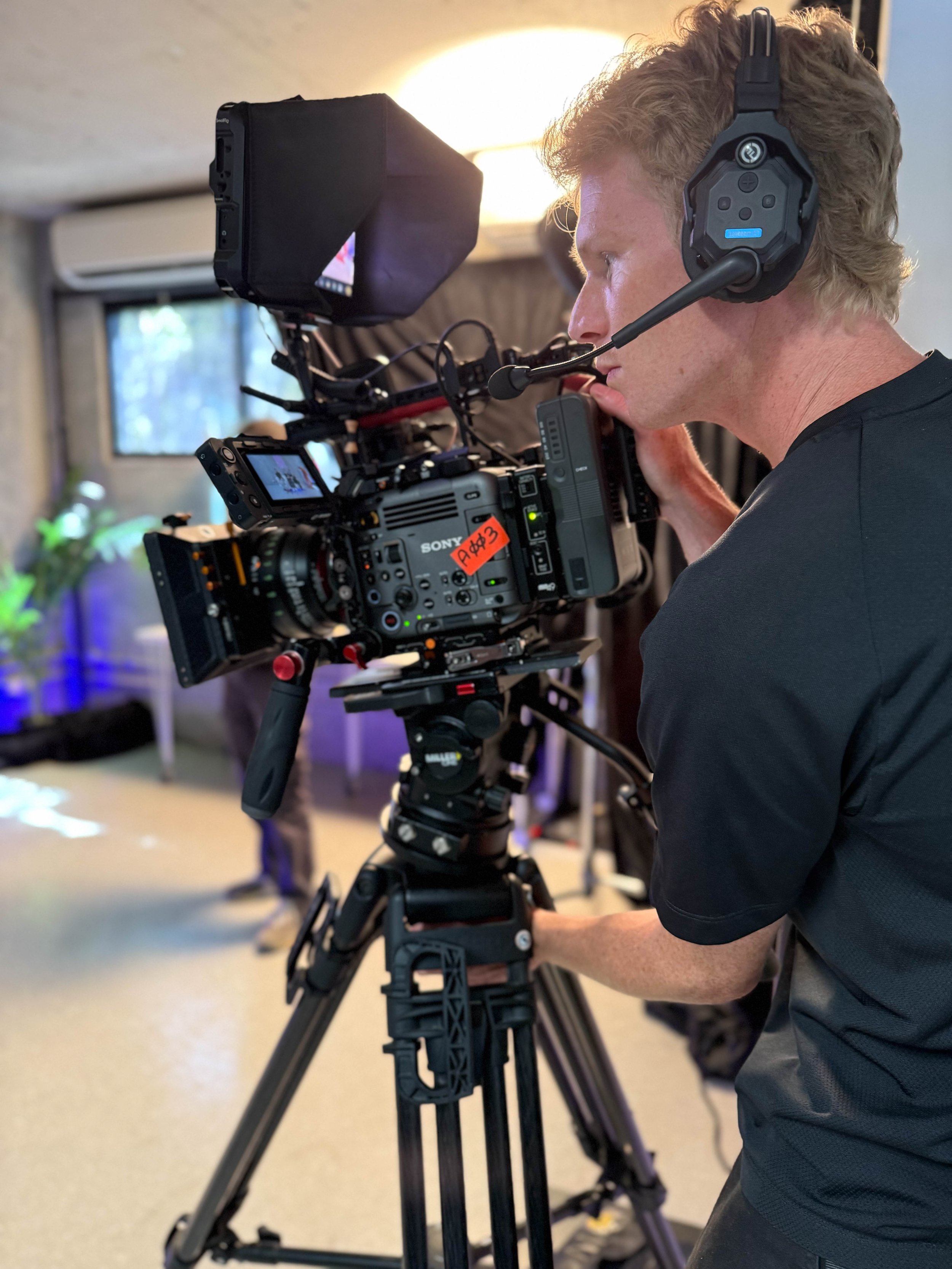 A man with curly blond hair wearing a black shirt using professional video camera equipment on a tripod, with a headset and microphone, in an indoor setting with a window and blurred background.