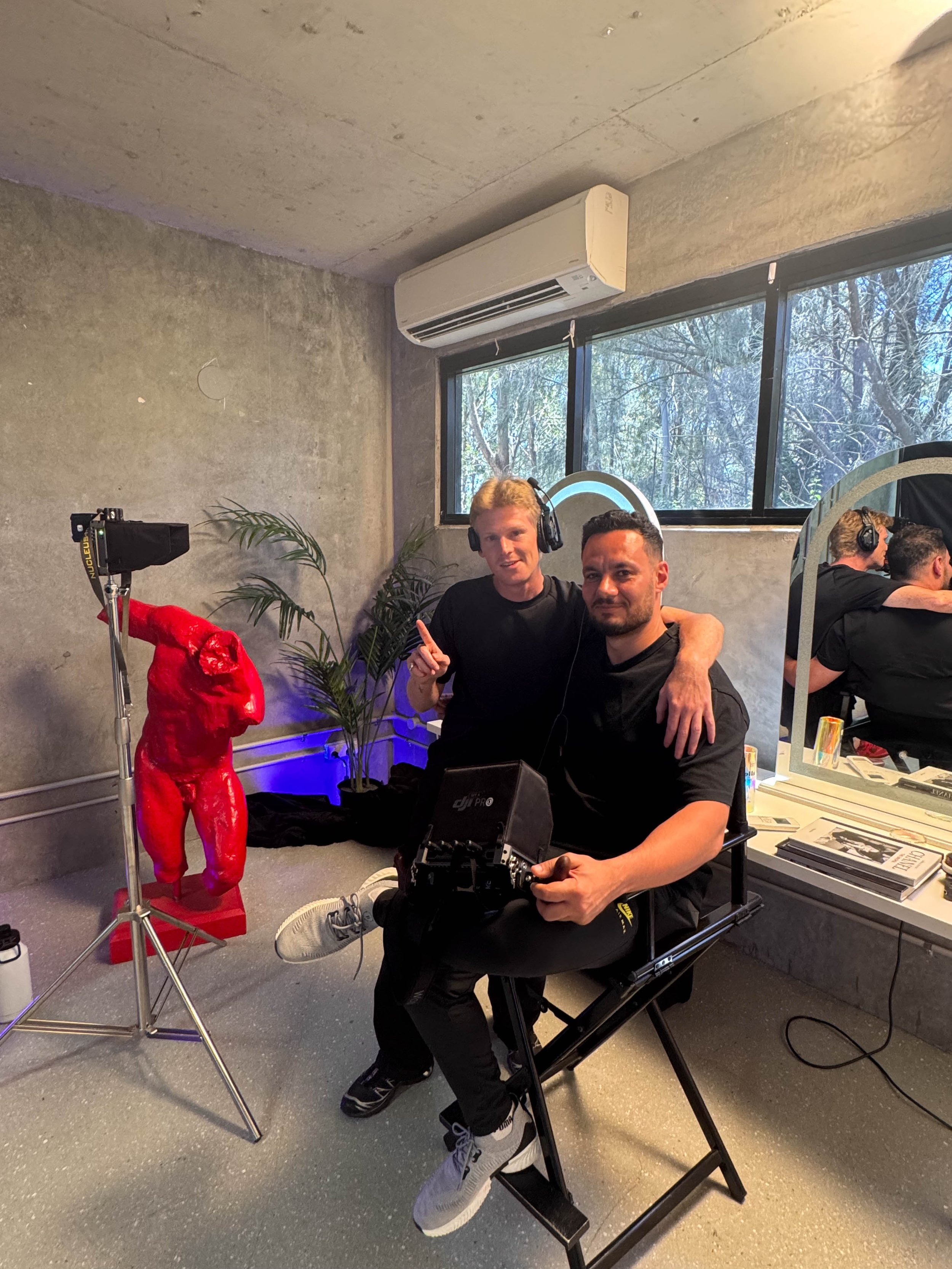 Two men sitting in front of a mirror in a room with a concrete wall, with DJ equipment and a red mannequin on a stand nearby. One is holding a camera stabilizer, while the other is smiling and making a pointing gesture.