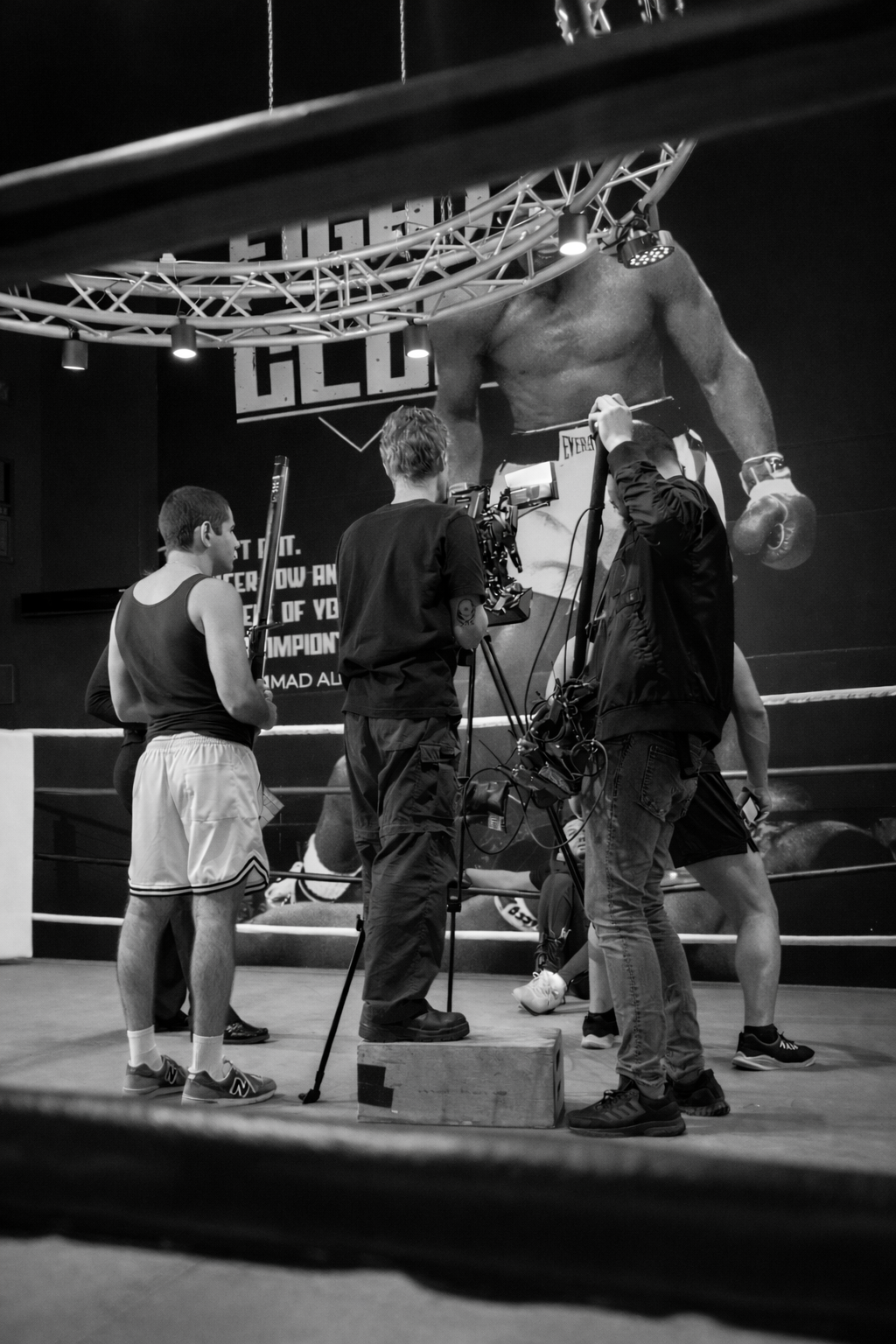 Behind-the-scenes photograph of a boxing gym, showing a filming crew setting up a shot with a large poster of a boxer on the wall in the background.
