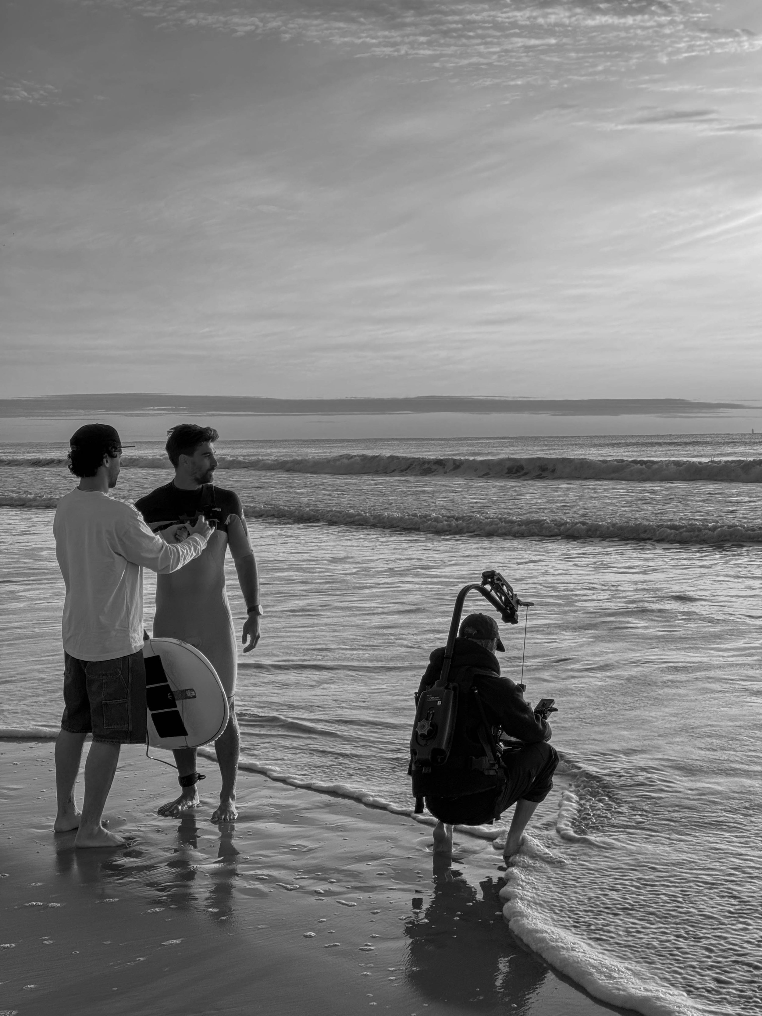 Two men are walking along the shoreline of a beach, one of them holding a surfboard, with a third person crouching with a camera and equipment, all near the ocean waves, under a partly cloudy sky.