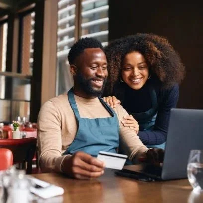 A happy couple shopping online together at a restaurant table, with a laptop and credit card.