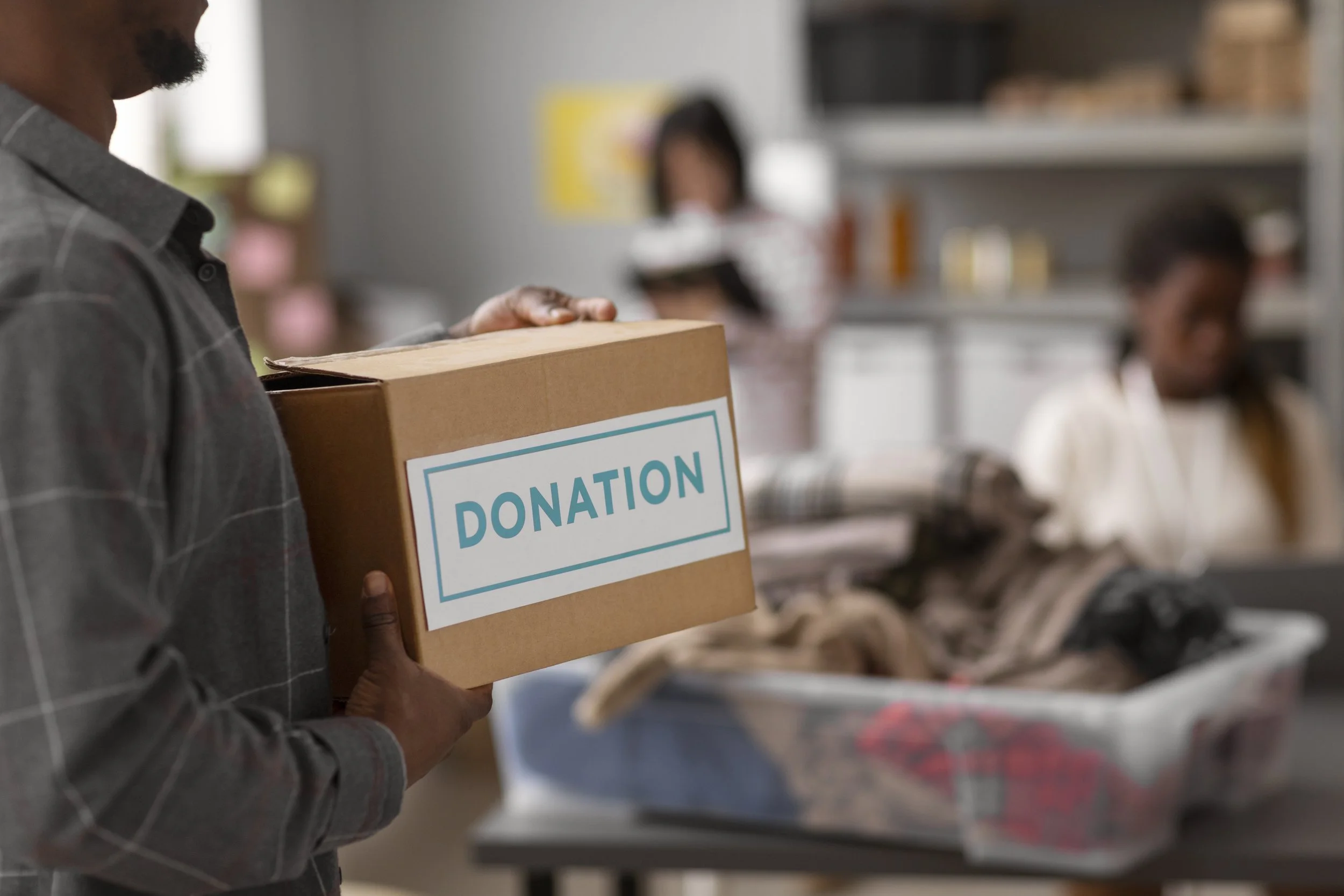 A person holding a donation box labeled 'DONATION' in a room with shelves and other people in the background.