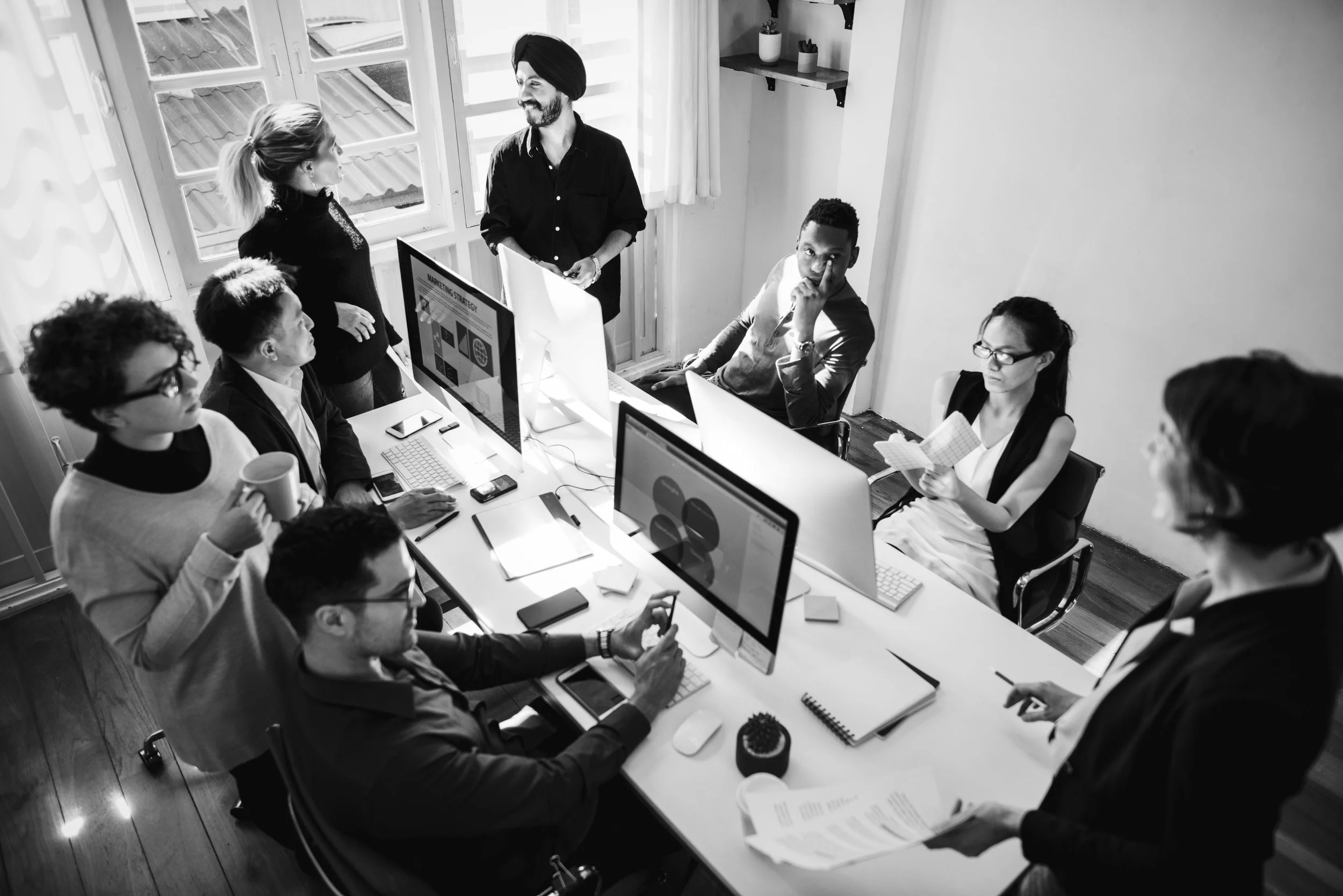A group of people in a meeting room with computers, some sitting and some standing, engaging in a discussion. One person appears to be presenting information on a screen.
