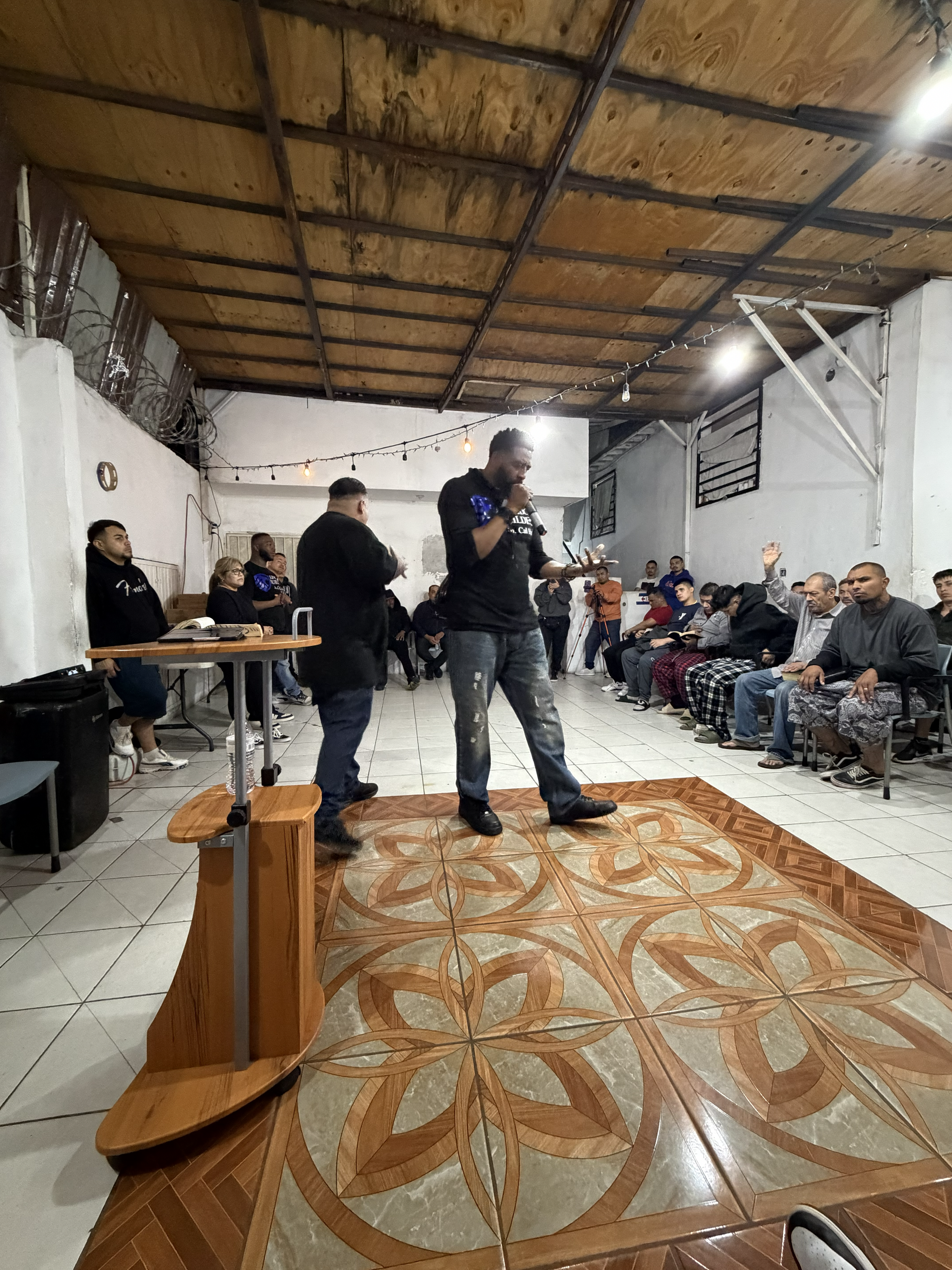 A man holding a microphone speaking in front of an audience seated on chairs inside a room with wooden ceiling panels, white painted walls, and decorative tile flooring.