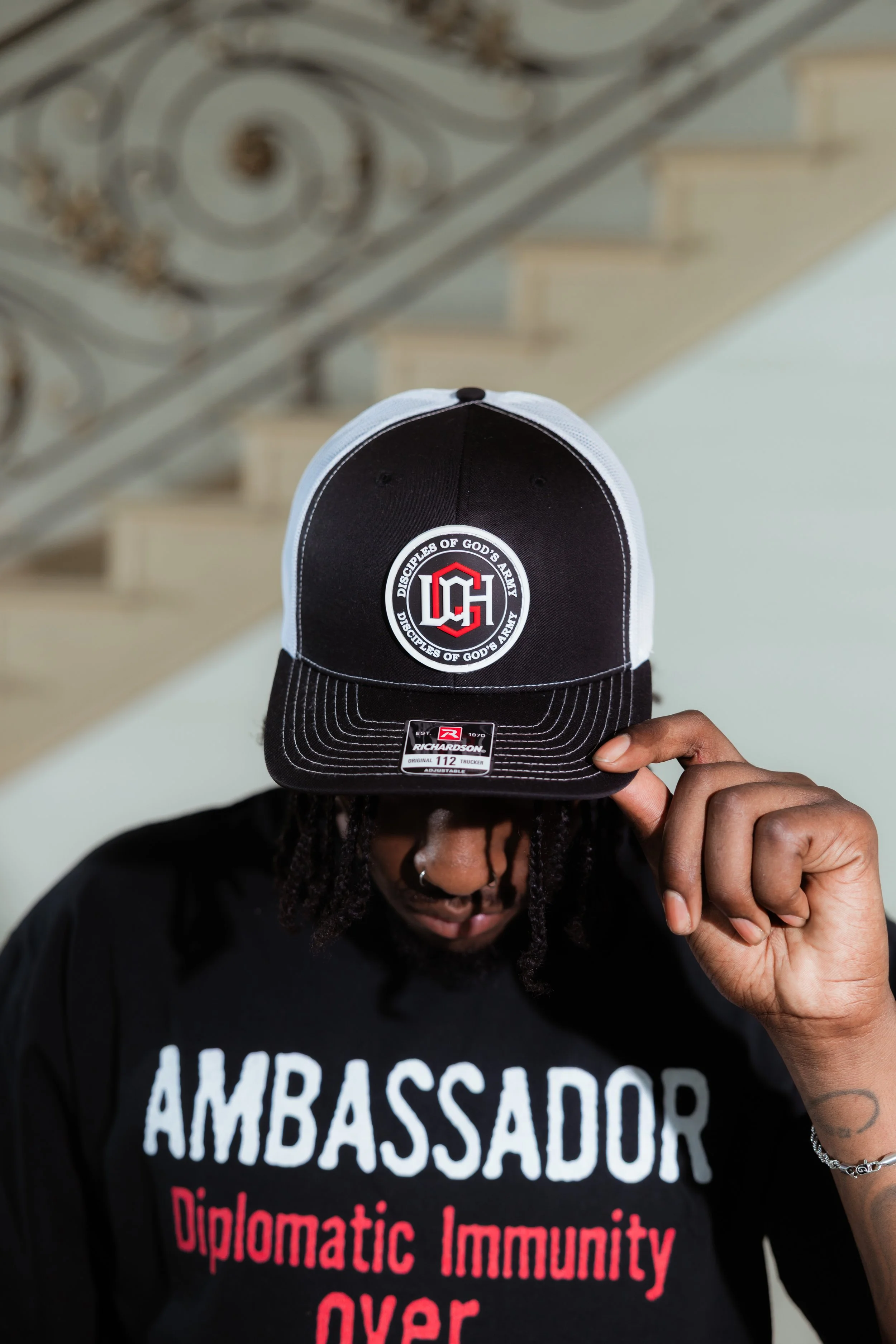 Person wearing a black baseball cap with a logo and text, holding the cap brim with their hand, wearing a black T-shirt with white and red text, standing indoors under a staircase with a decorative metal railing.