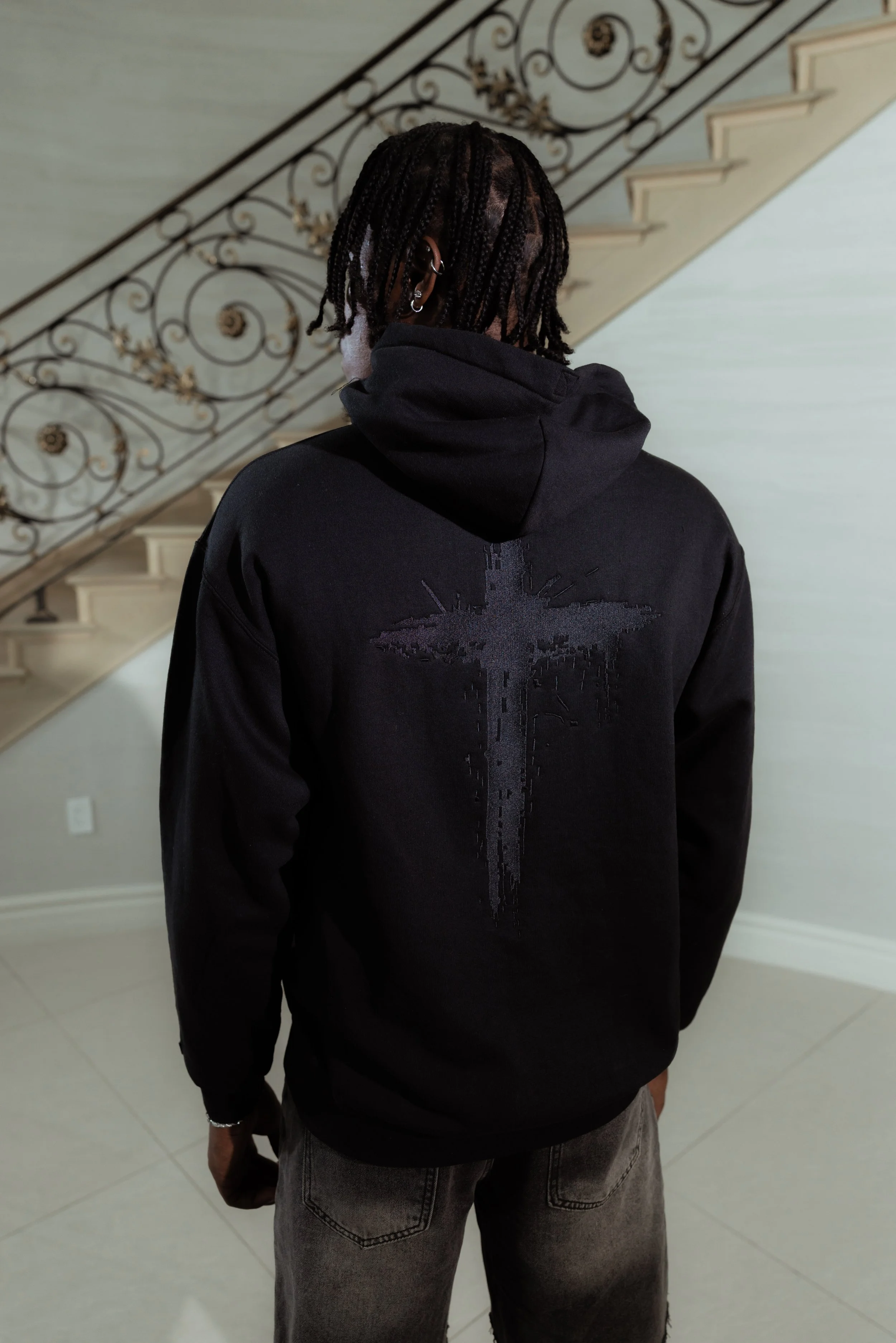 Person wearing a black hoodie with a distressed cross design on the back, gray jeans, and earrings, standing indoors near a staircase with ornate iron railing.