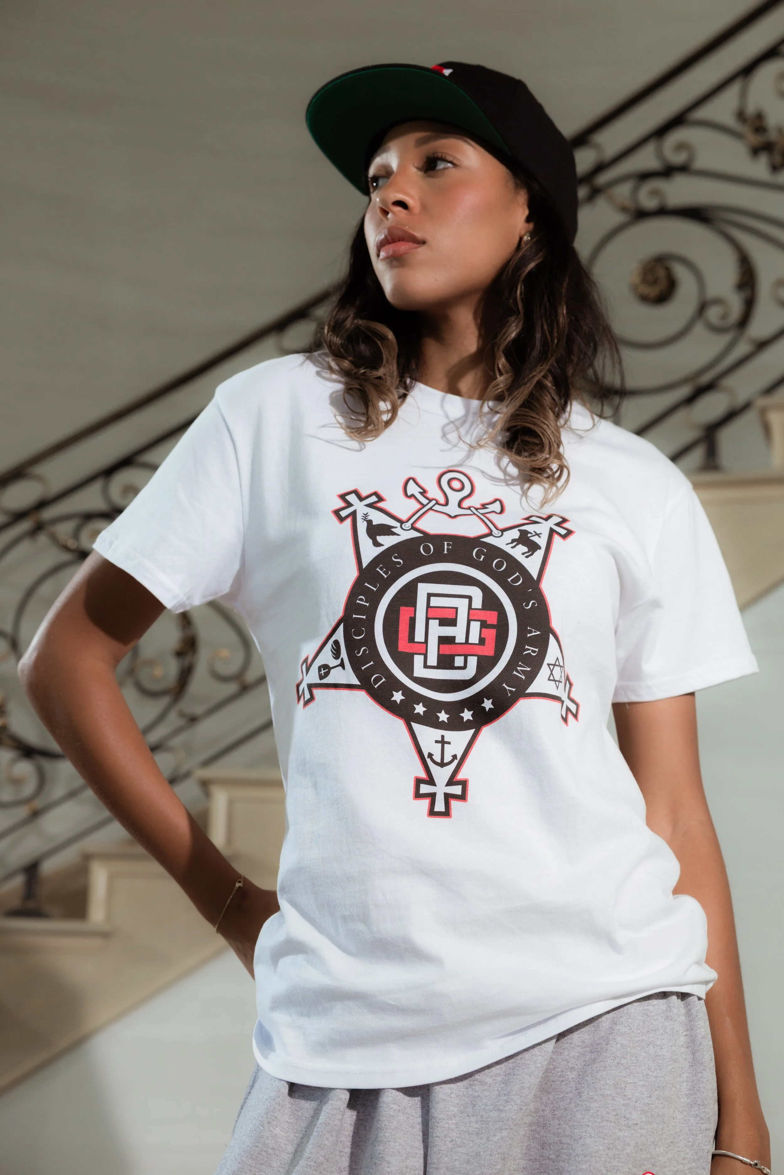 A woman wearing a black baseball cap and white t-shirt with a theological emblem, standing indoors with a staircase and ornate railing in the background.