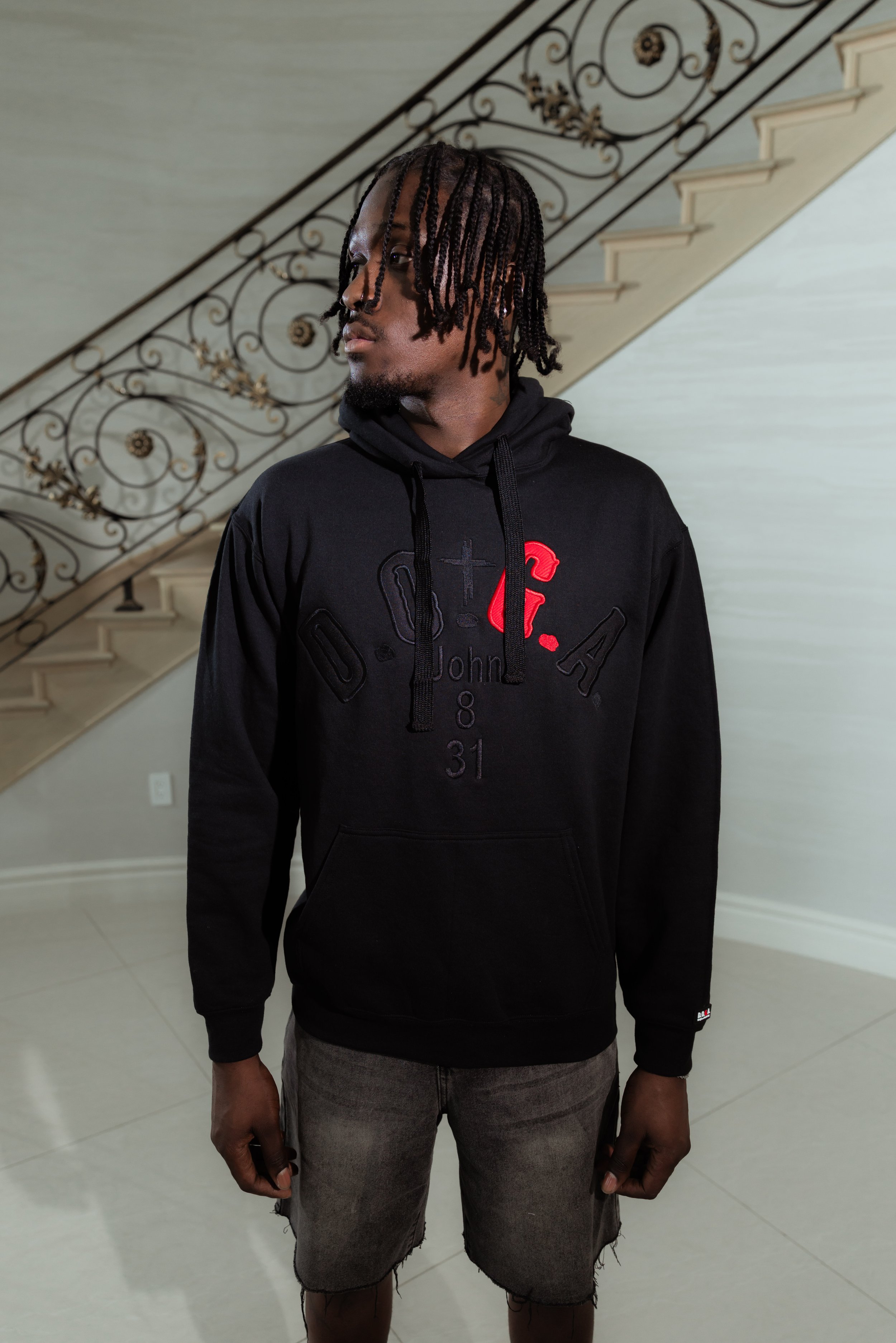 A young man with dreadlocks wearing a black hoodie with red and black text, gray shorts, standing indoors near a staircase with ornate railing, looking to his left.