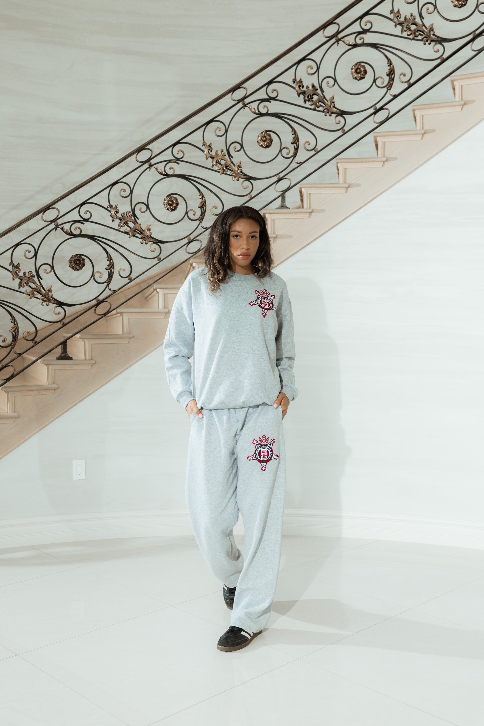 Young woman standing indoors near a staircase with ornate black metal railings, wearing a casual light gray sweatshirt and sweatpants with a red and white emblem, and black sneakers.