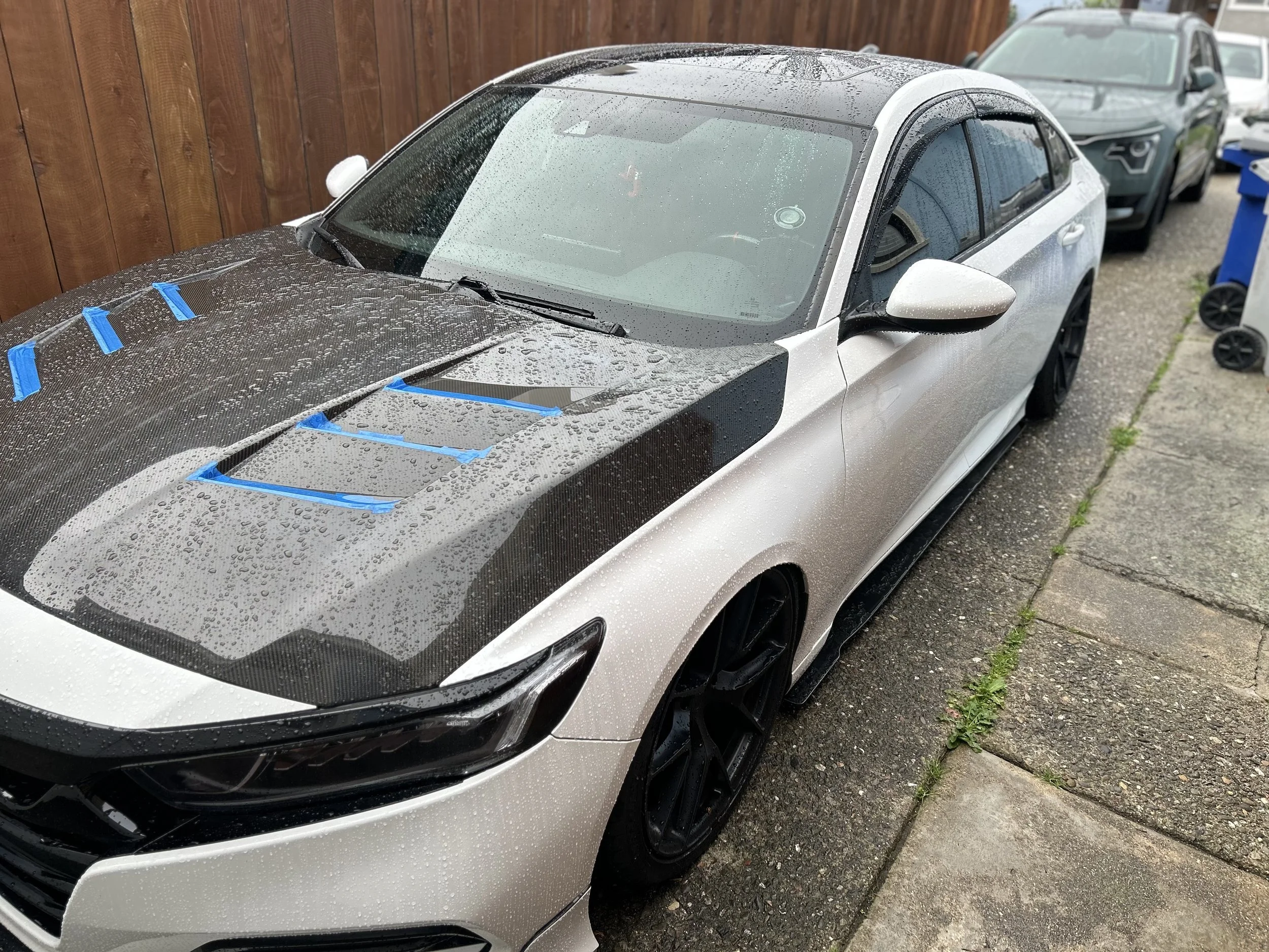 A silver and black sports car parked outdoors next to a wooden fence, with raindrops on its surface and blue painter's tape on the hood and roof. Several other vehicles are visible parked nearby.