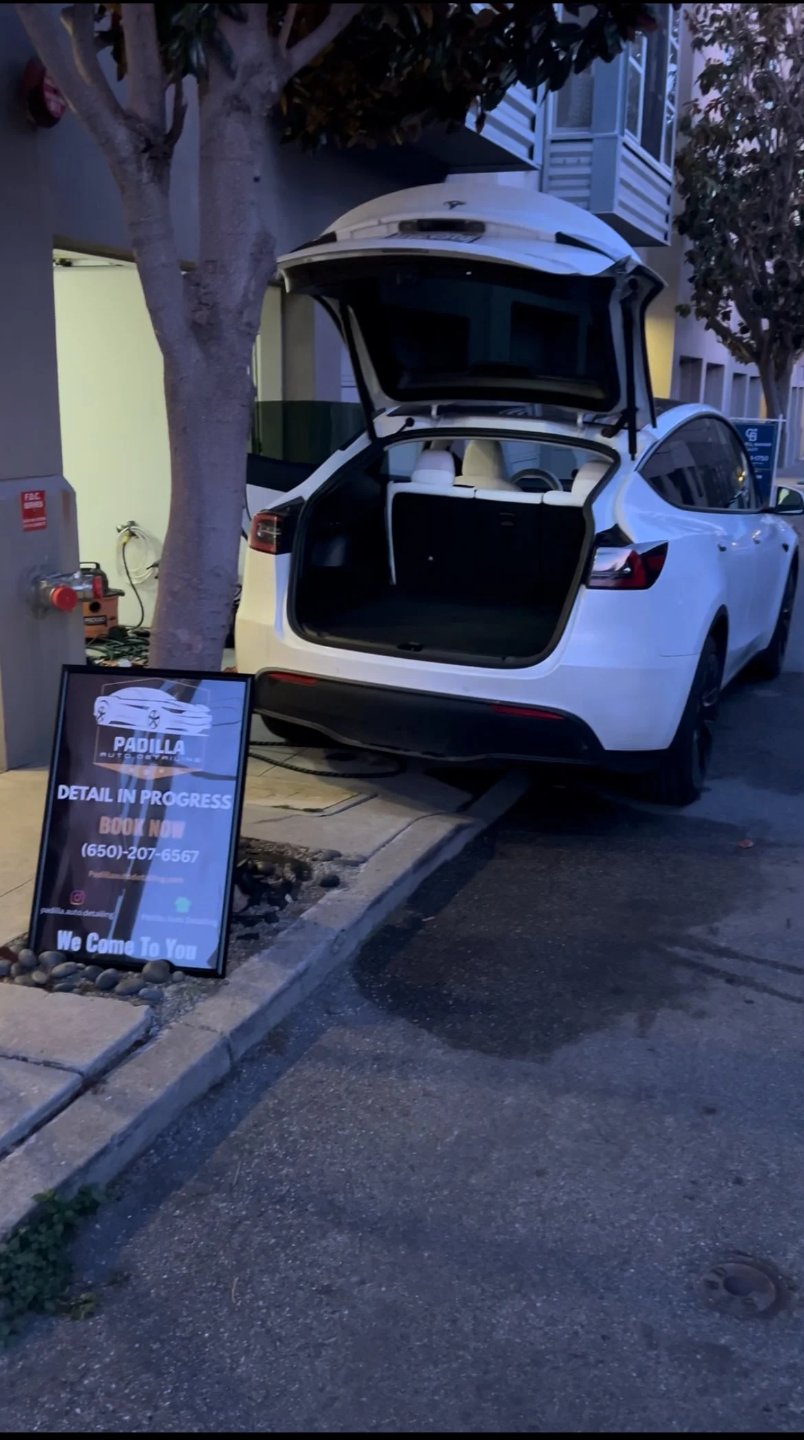 A white electric vehicle with stock open, parked on the street next to a sidewalk. There's a sign on the sidewalk advertising a car detail service, and a tree partially blocking the view of the vehicle.