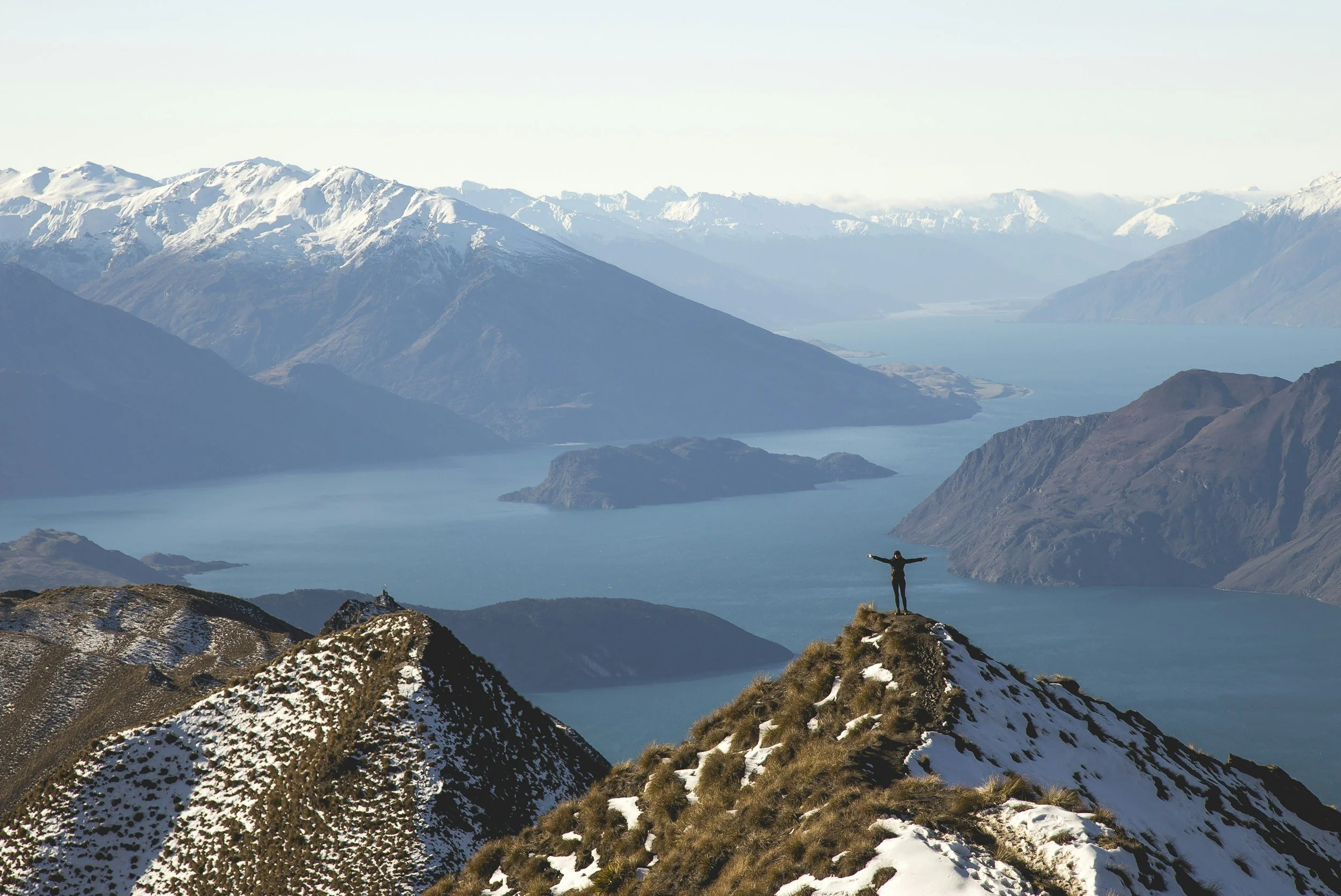 Person standing with arms outstretched on mountain peak overlooking snow-capped mountains and a lake.