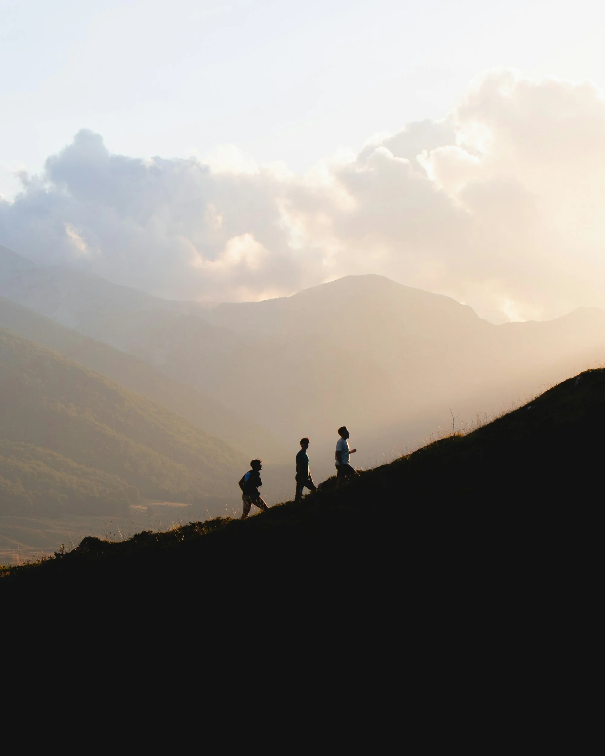 Three people hiking up a grassy hill during sunset with mountains and clouds in the background.