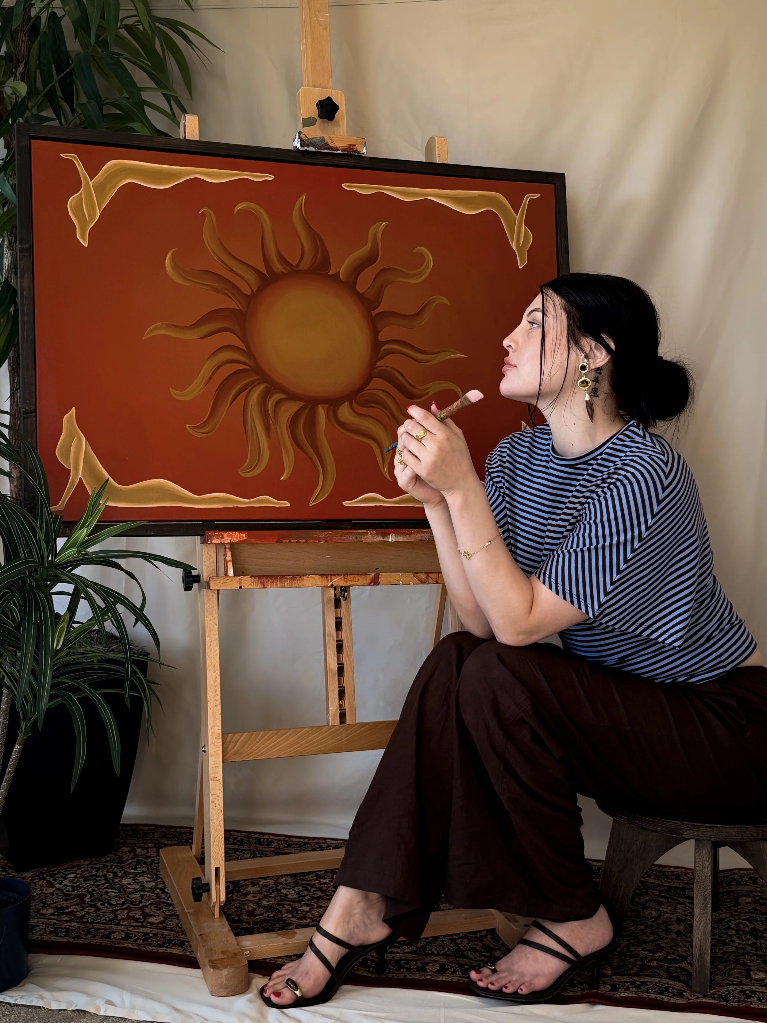 A woman sitting in front of an easel with a painting of a sun, wearing a striped shirt, black pants, and black strappy sandals, holding a large paintbrush.