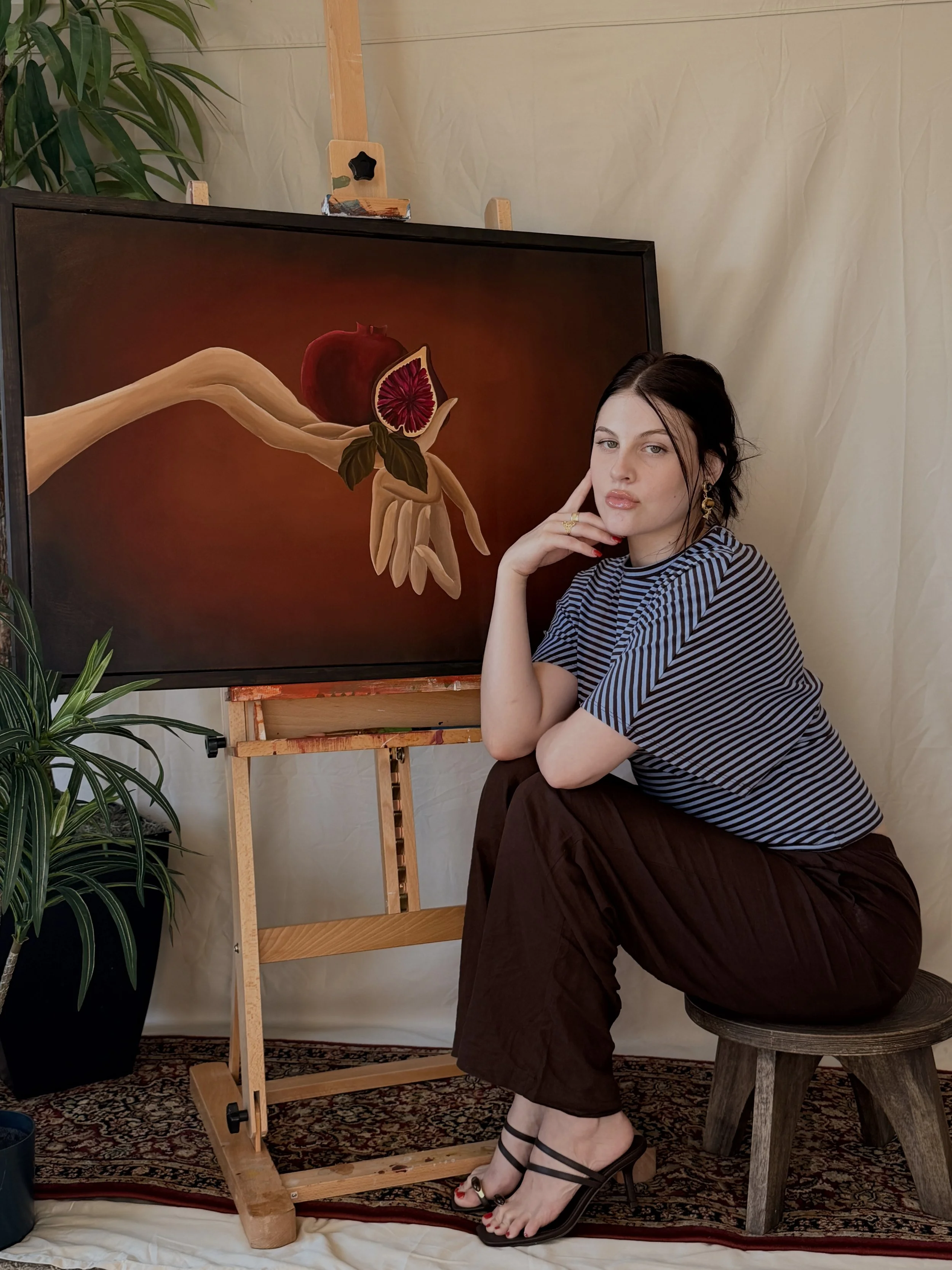 A woman with dark hair, wearing a striped shirt and brown pants, sitting on a stool in front of a painting of a hand holding an pomegrante with leaves, with plants and a lamp in the background.