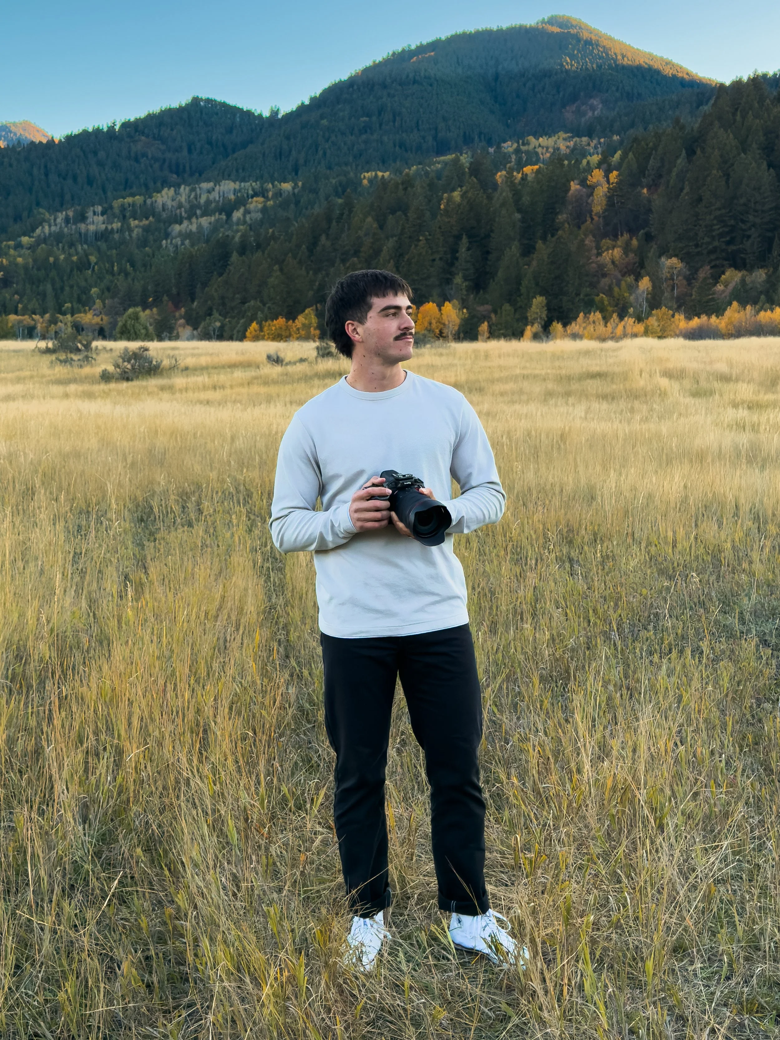 A young man with dark hair and a mustache stands in a grassy field holding a professional camera. He wears a light gray long-sleeve shirt, black pants, and white sneakers. Behind him, there are rolling hills or mountains covered with dense trees, with some autumn-colored foliage, under a clear blue sky.