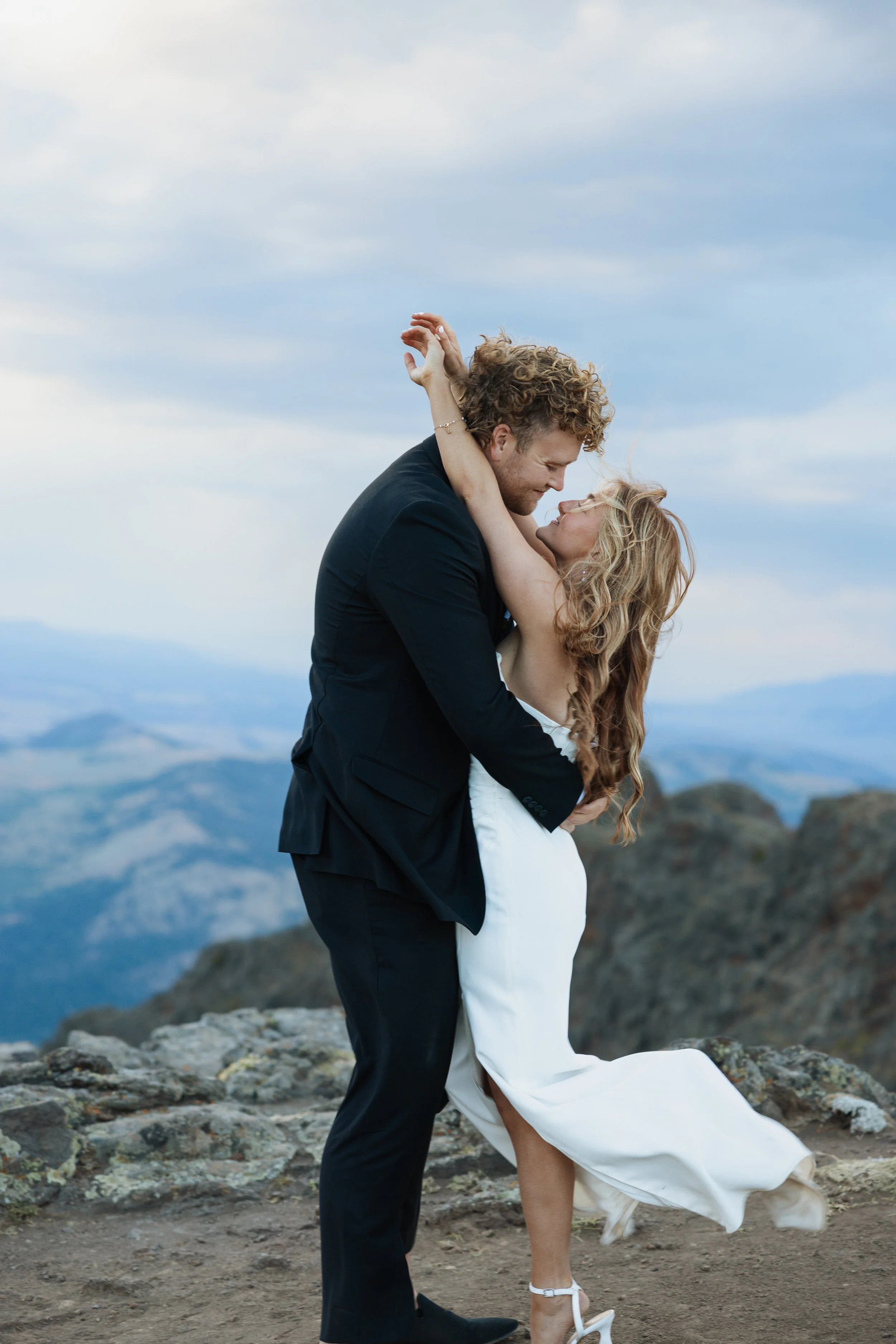 A couple dancing on a rocky mountaintop with mountain ranges in the background, woman in a white dress and heels, man in a black suit, embracing and smiling.