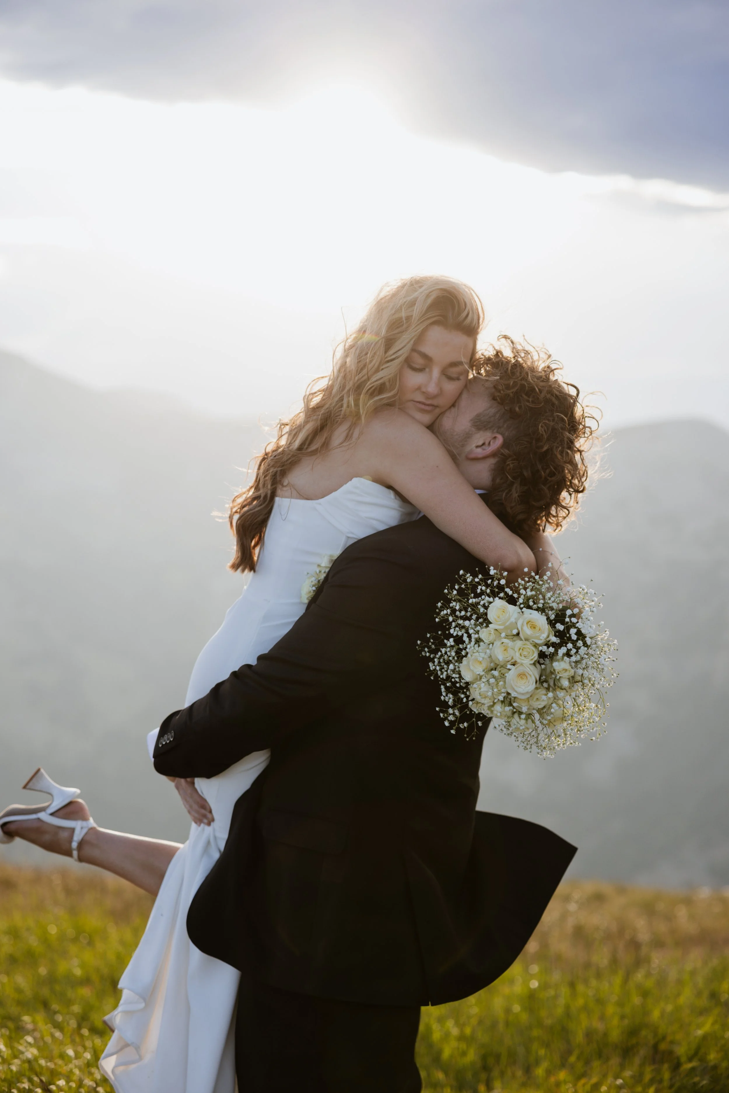 A man in a black suit lifting a woman in a white wedding dress in a grassy outdoor setting during sunset, with mountains in the background, holding a bouquet of white roses and baby's breath.