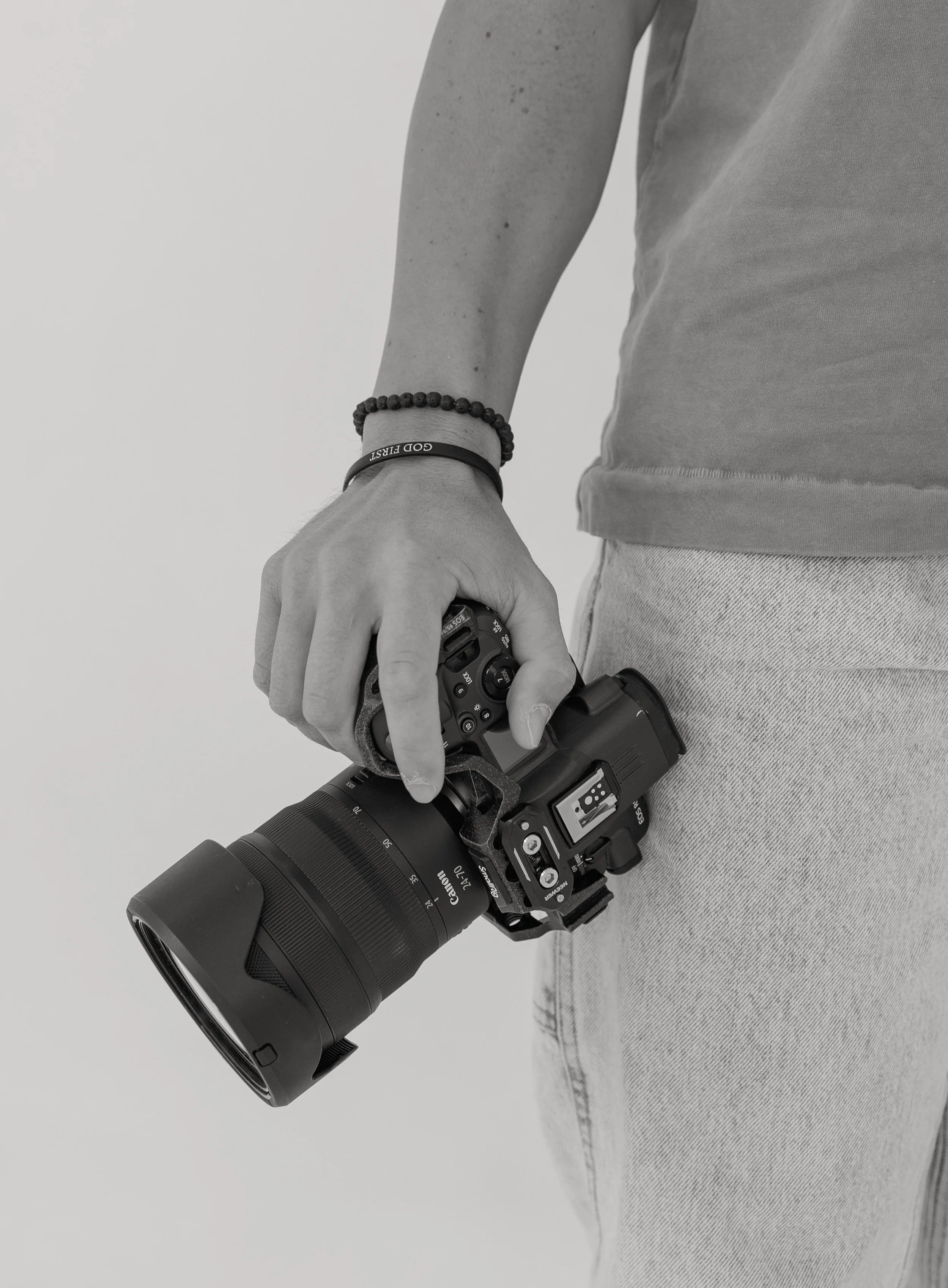 A person holding a professional camera with an attached telephoto lens, wearing a bracelet and a beaded wristband on their left wrist, dressed in a t-shirt and light-colored jeans.
