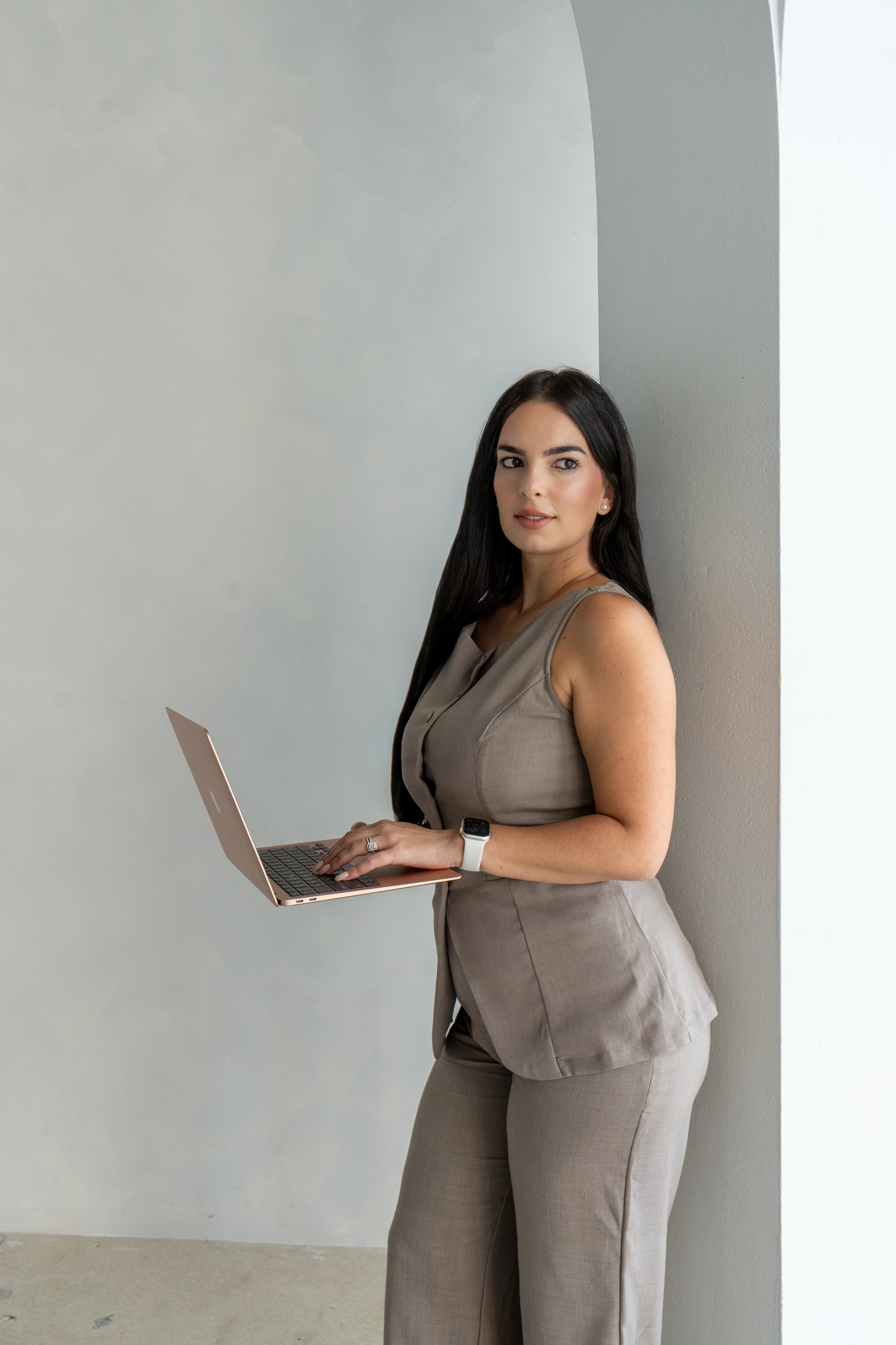 A woman with long dark hair, wearing a beige sleeveless suit and a smartwatch, leaning against a wall while holding a rose gold laptop.