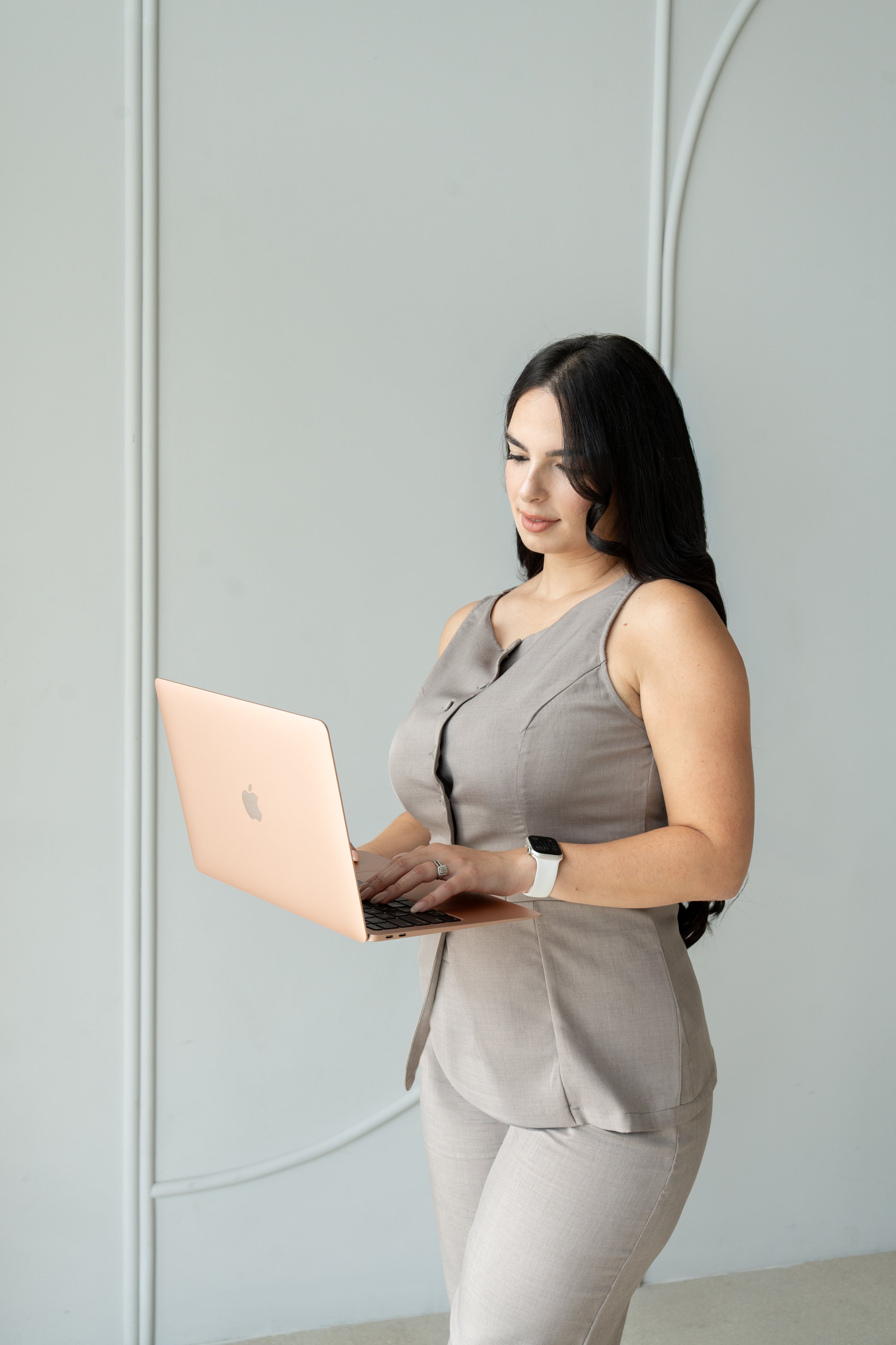 A woman wearing a beige sleeveless suit holding a pink MacBook laptop, working in a modern interior with light-colored walls.