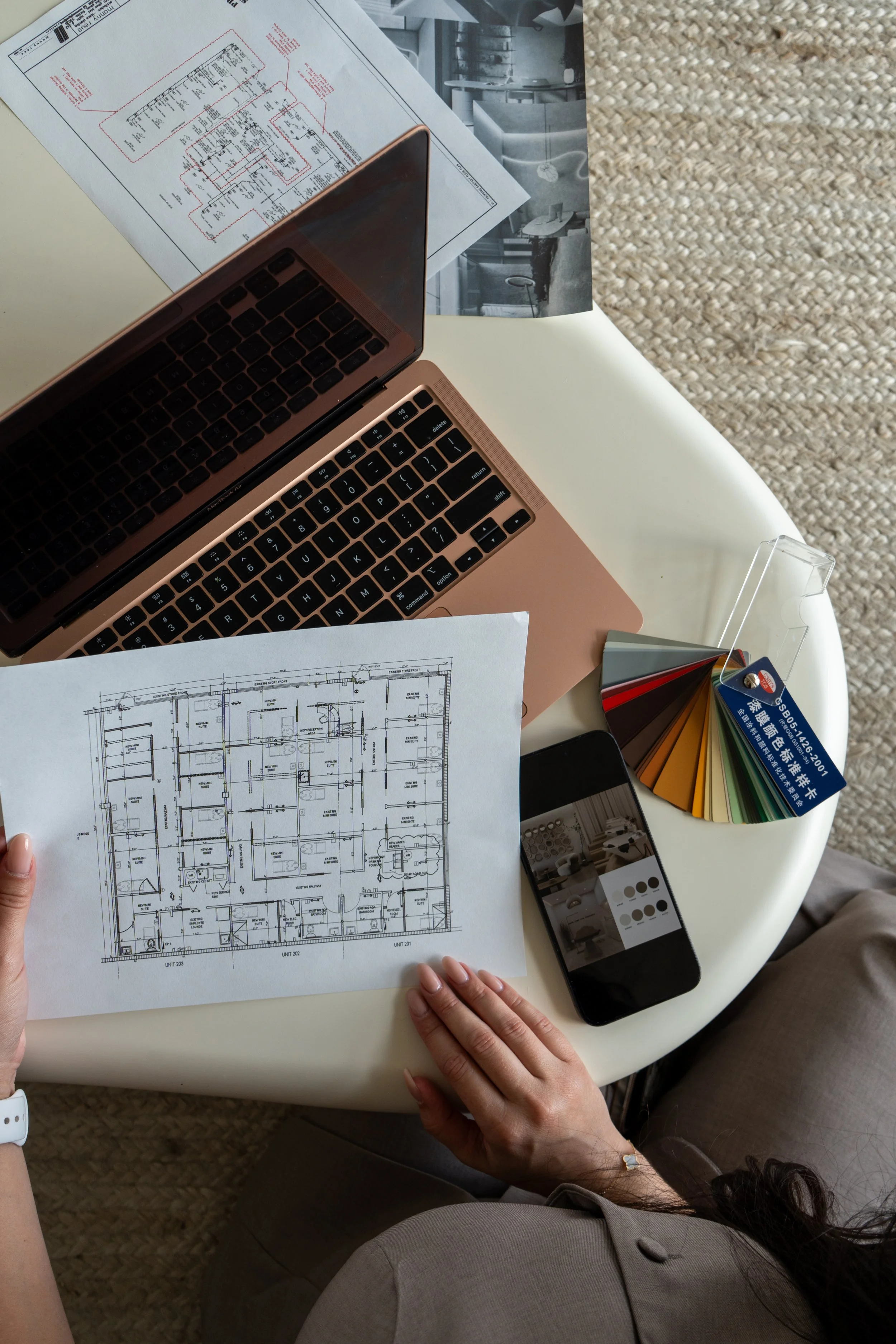 Person holding architectural blueprint with a laptop, color swatches, smartphone, and printed plans on a white table.
