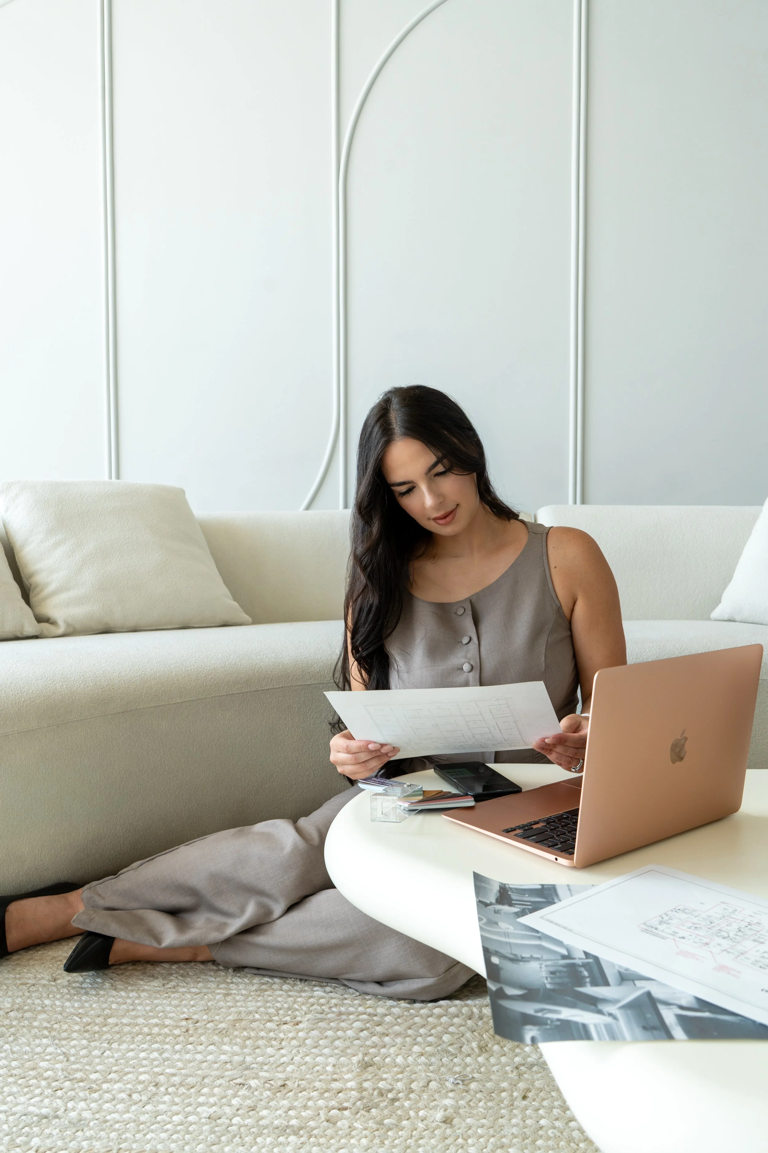 A woman sitting on the floor with her legs crossed, looking at printed documents with a laptop open on a round table in front of her. There are some photographs and papers on the table.