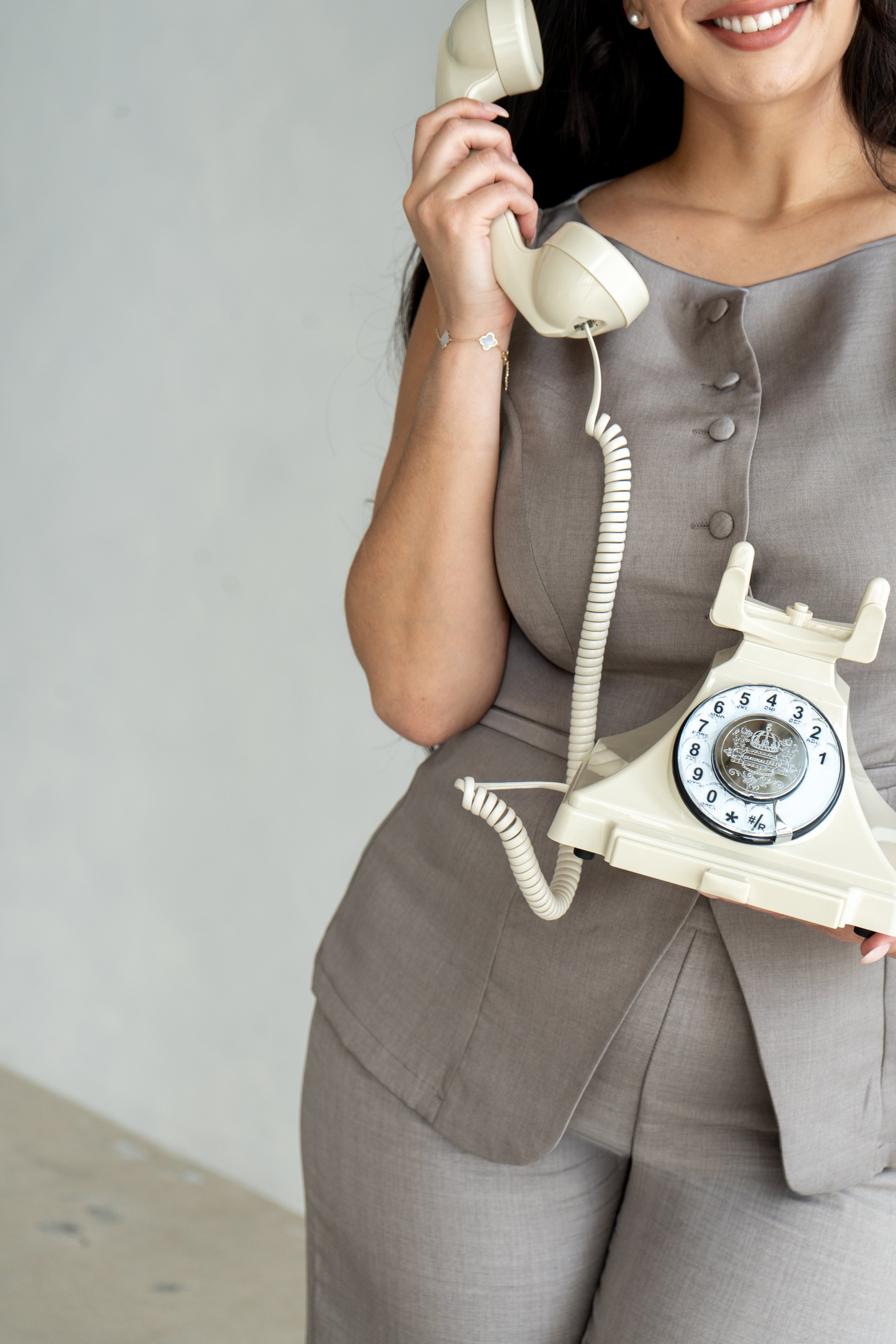 A woman in a beige suit holding a vintage beige rotary phone, smiling.