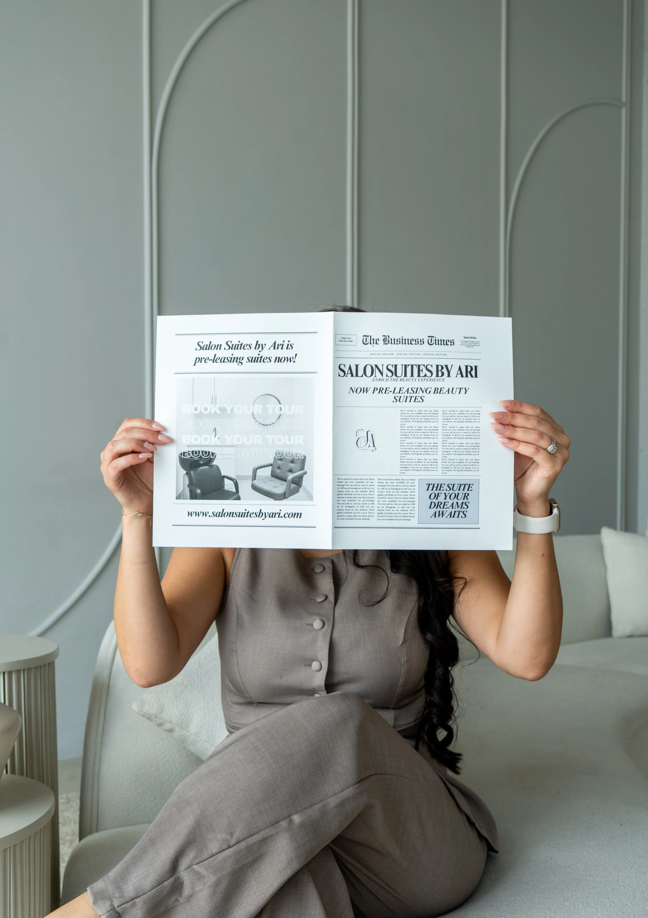 A woman sitting on a cream-colored couch reads a newspaper promoting Salon Suites by Ari. She is wearing a beige sleeveless jumpsuit, has long dark hair, wears a watch and ring, and is in a modern, neutral-toned room with a small side table.